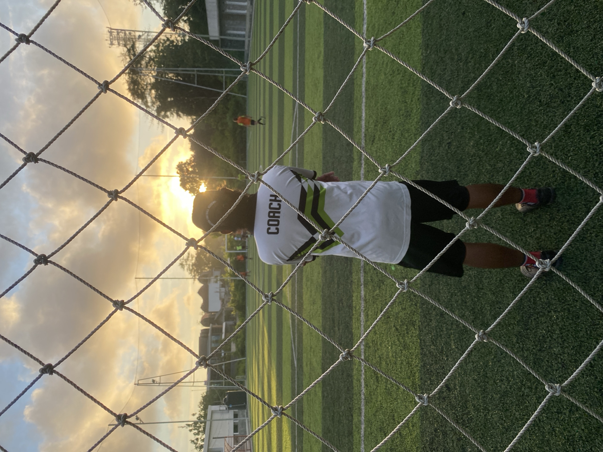 The image shows a person wearing a white t-shirt with "COACH" printed on the back. They are standing behind a net fence, looking towards a soccer field at sunset. The field has green artificial turf with white lines. In the background, other people can be seen playing on the field. Some buildings are visible in the far background, along with trees and a partly cloudy sky. The photo appears to have been taken in Kuta Selatan, Indonesia.