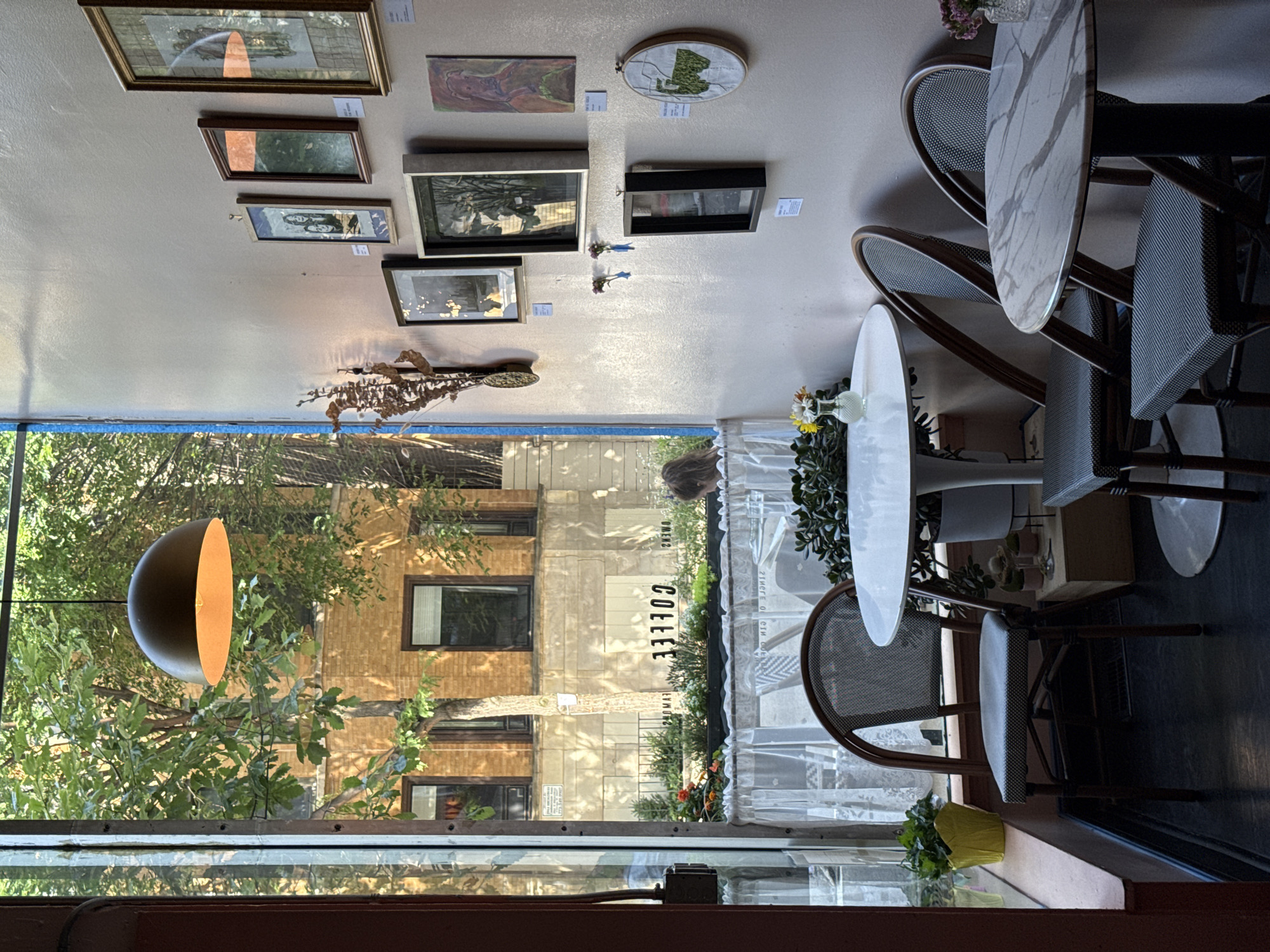 This image depicts the interior of a cafe, likely in Chicago, USA, viewed through a large window that looks out onto a street scene. Inside the cafe, several small tables are set with chairs. The decor includes various framed artworks displayed on a light pink wall, and a hanging pendant light fixture.
Looking through the window, a brick building with multiple windows is visible across the street. There are trees with green foliage in the foreground, casting shadows. A sign with the word "COFFEE" in black lettering is visible on the exterior of the building across the street. Some planters with greenery and flowers are also seen outside.
The lighting suggests it is daytime, with sunlight filtering into the cafe, creating bright and shaded areas. A person with dark hair is partially visible through the window, seated at one of the tables inside the cafe across the street. The overall atmosphere appears to be relaxed and inviting.