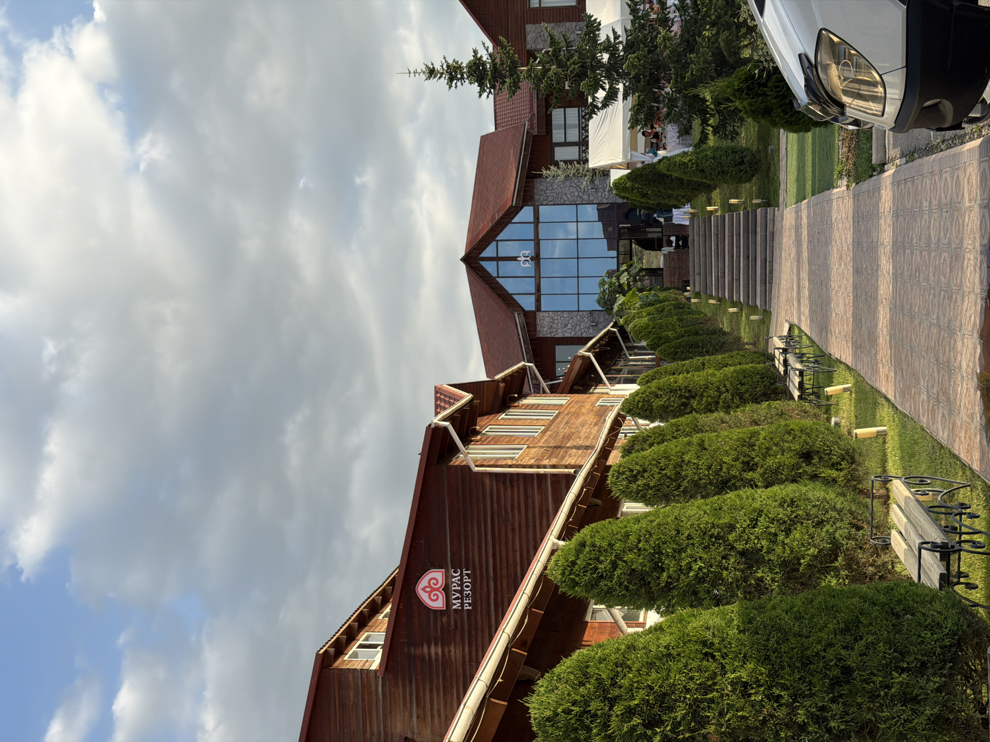 This image captures a view of the "Muras Resort" building in Bishkek City, Kyrgyzstan. The building is primarily constructed of dark wood, with a distinctive red-tiled roof and a large, triangular glass facade. The resort's logo, a stylized heart-like symbol with text in Cyrillic that reads "МУРАС РЕЗОРТ", is visible on the wooden exterior.
The scene is outdoors, with a pathway made of patterned tiles leading up to the building. Flanking the pathway are meticulously manicured green bushes arranged in a row, creating a formal garden effect. Beyond the bushes, a well-maintained lawn stretches out. In the background, several trees, including a tall evergreen, are visible, along with portions of a white tent structure. A white vehicle is parked on the right side of the frame, near the pathway.
The sky is filled with a mix of white and gray clouds, suggesting it might be late afternoon or early evening. The lighting indicates soft sunlight filtering through the clouds, casting gentle shadows. There are no people prominently featured in the image, but a few figures can be faintly seen under the tent structure in the distance. The overall impression is one of a well-kept and serene resort setting.