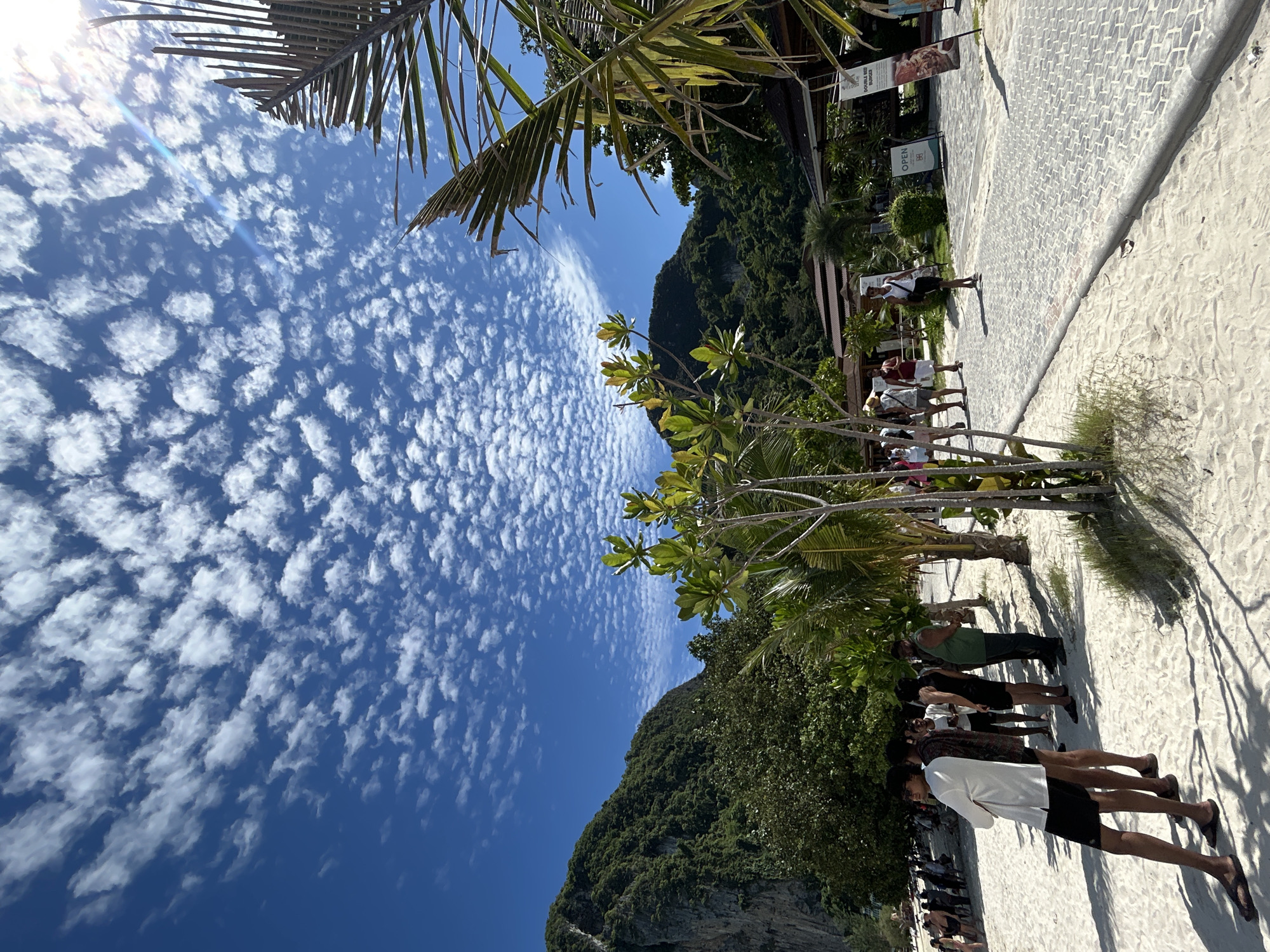 The image captures a tropical beach scene in Ao Nang, Thailand, characterized by a vibrant blue sky filled with scattered white, puffy clouds. In the foreground, white sand stretches across the beach, casting long shadows from the sparse vegetation. Several individuals are present, some standing in small groups, while others walk along a paved pathway that borders the sand. These individuals are dressed in casual, summer attire.
To the left of the frame, lush green mountains rise majestically, densely covered with trees and vegetation, suggesting a mountainous terrain typical of the region. Palm trees and other tropical foliage are scattered throughout the scene, adding to the exotic atmosphere. In the background, several buildings with tiled roofs are visible, hinting at the presence of shops or restaurants. One sign on a building clearly reads "OPEN." The overall impression is one of a popular tourist destination, bathed in bright sunlight.