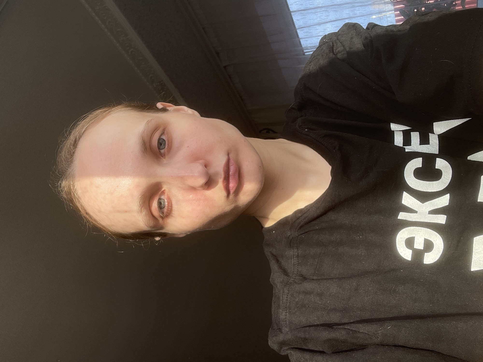The image, rotated 90 degrees clockwise, features a young woman from Kuyeda, Russia, captured in a close-up, likely a selfie. She is illuminated by strong natural light, creating a dramatic contrast across her face. Her head is tilted, and her light blue or grey eyes gaze directly at the camera. Her light-colored hair is pulled back from her face, which displays a neutral, calm expression.
The right side of her face and shoulder are brightly lit by direct sunlight, while the left side is in deep shadow. She is wearing a black t-shirt made of a soft fabric, which shows some visible lint or dust. Large white Cyrillic letters are printed on the shirt; the visible portion clearly spells "ЭКСЕ," likely part of a longer word such as "ЭКСЕЛЕНТ" (Excellent).
The background is predominantly dark and out of focus. In the upper right corner of the original orientation (which is the current top-right after rotation), a sliver of a sunlit window with what appears to be blinds or curtains is visible, suggesting an indoor setting during the daytime.