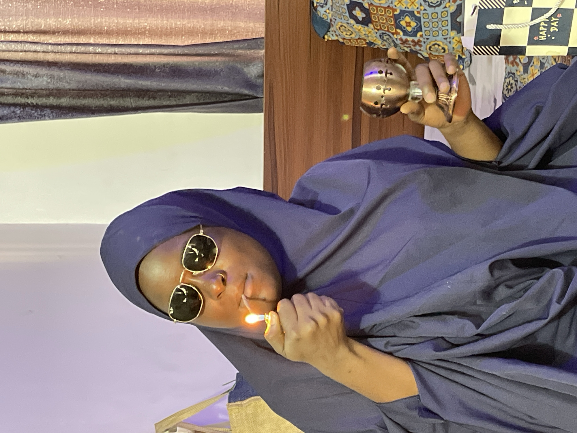 A person wearing sunglasses and a blue hijab is holding a lit lighter in one hand and a metal incense burner in the other. The location is Gwarinpa, Nigeria.