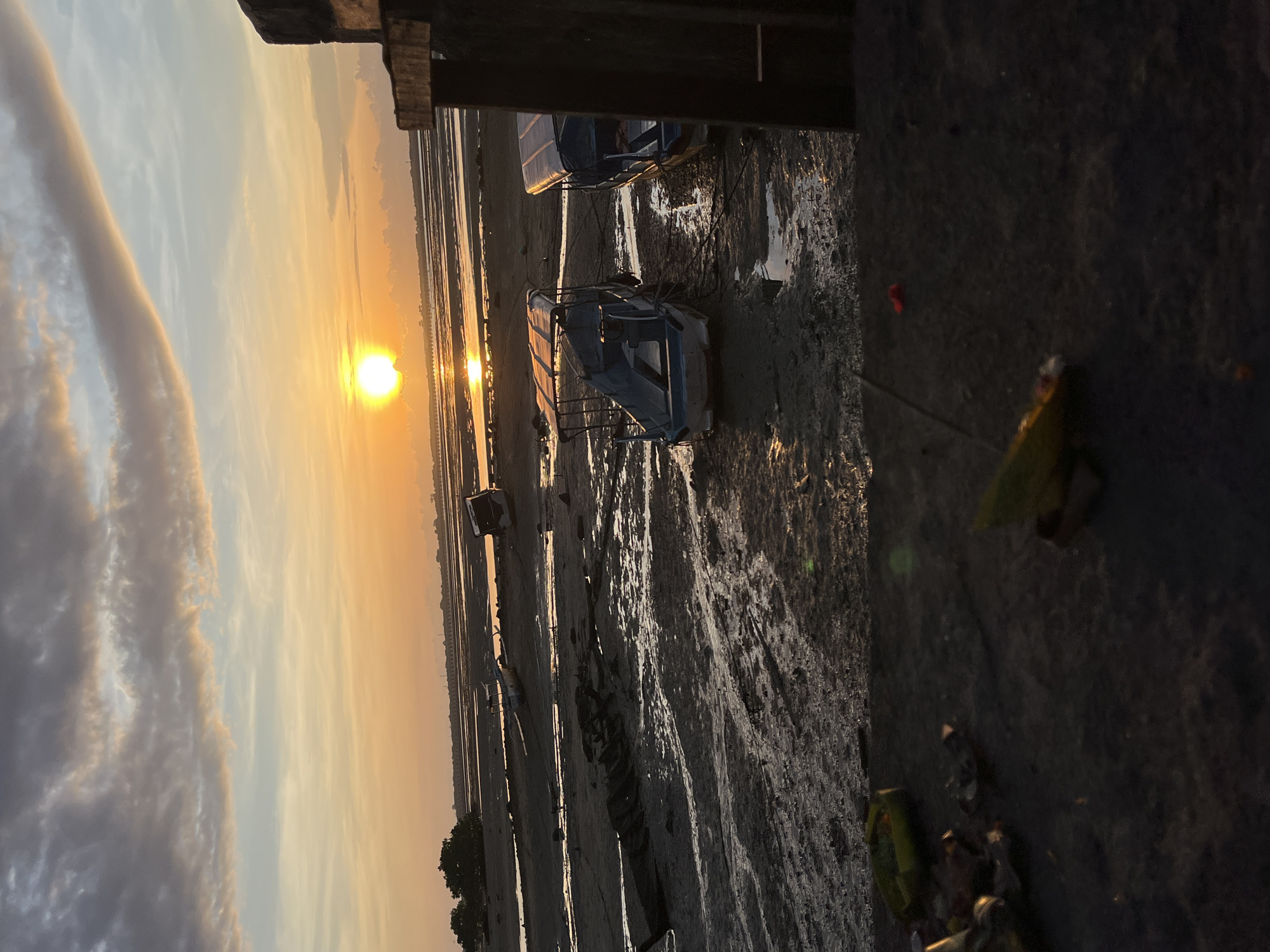 The image, taken at sunset, depicts a wide, muddy tidal flat in what appears to be a coastal area, likely in Kuta Selatan, Indonesia. The sun is a bright, luminous orb, low on the horizon, casting a warm golden and orange glow across the sky and reflecting intensely on the wet surfaces of the distant water and the exposed mud flats. The sky transitions from vibrant yellow and orange near the sun to softer blues and grays, with a distinct, elongated cloud formation stretching across the upper sky.
In the mid-ground, several small, light-colored boats, possibly traditional fishing or tour boats, are beached on the muddy terrain, indicating a low tide. Ropes can be seen tethering some of these vessels. The foreground on the right side of the image features a dark, wooden structure, likely part of a pier or elevated platform, partially framing the scene. The muddy ground is textured and reflective, catching the golden light.
The scene is tranquil and serene, capturing the peaceful transition from day to evening. No people are visible in the image. There is no discernible text.