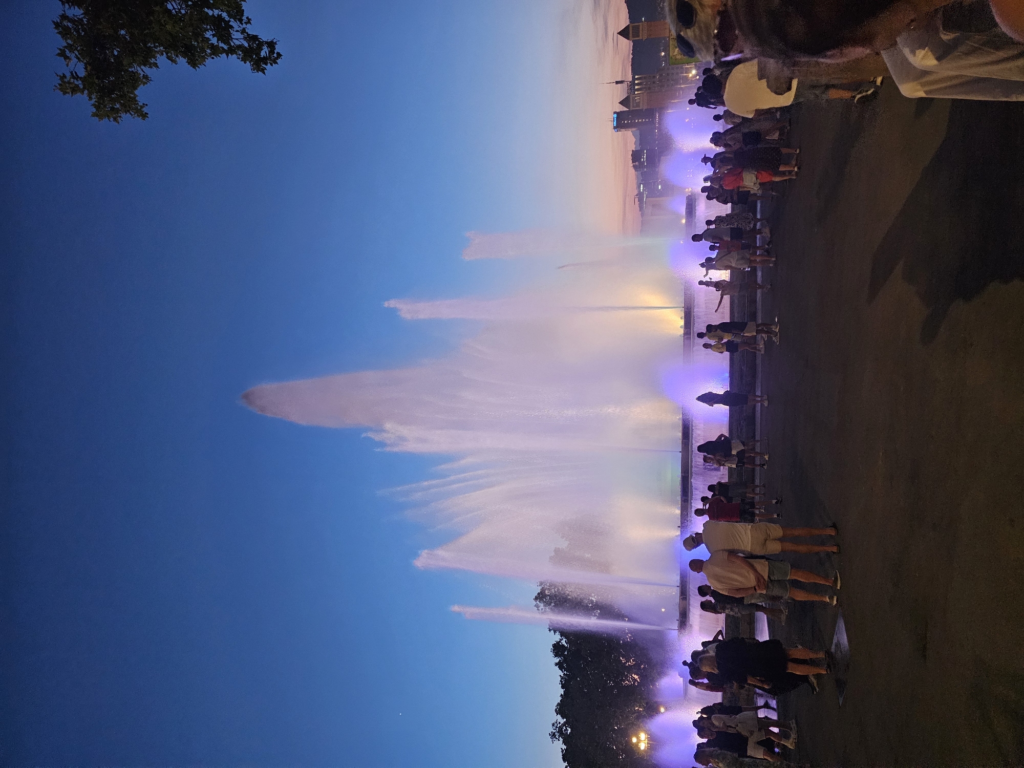 The image captures a nighttime view of the Magic Fountain of Montjuïc in Barcelona, Spain. A large, multi-tiered fountain is illuminated with vibrant purple and yellow lights, projecting plumes of water high into the clear blue evening sky. A crowd of people stands on the dark pavement in front of the fountain, observing the spectacle. The figures are mostly silhouetted against the bright fountain, with some individuals standing closer to the foreground. In the background, the city skyline of Barcelona is visible, with illuminated buildings and distant hills. The overall scene depicts a lively public gathering enjoying a nighttime light and water show.