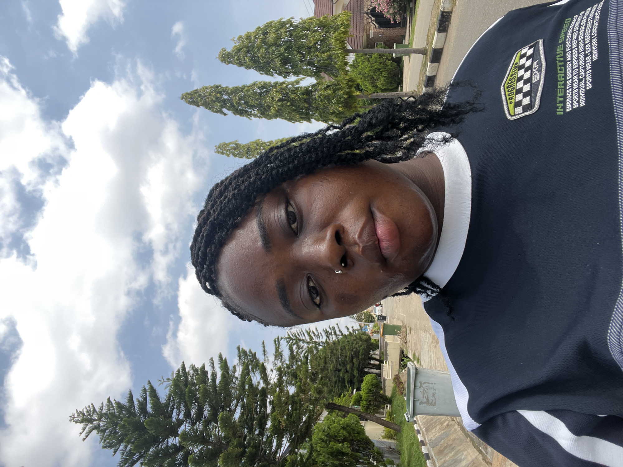 A young Black woman with braided hair is captured in a close-up, outdoor selfie. She is wearing a black and white long-sleeved shirt. The background suggests a suburban environment in Abuja, Nigeria, with several tall, slender trees and residential buildings visible. The sky is partly cloudy. The woman has a nose ring. To her left, a green trash bin is present on the side of a paved road. The overall lighting indicates daytime.