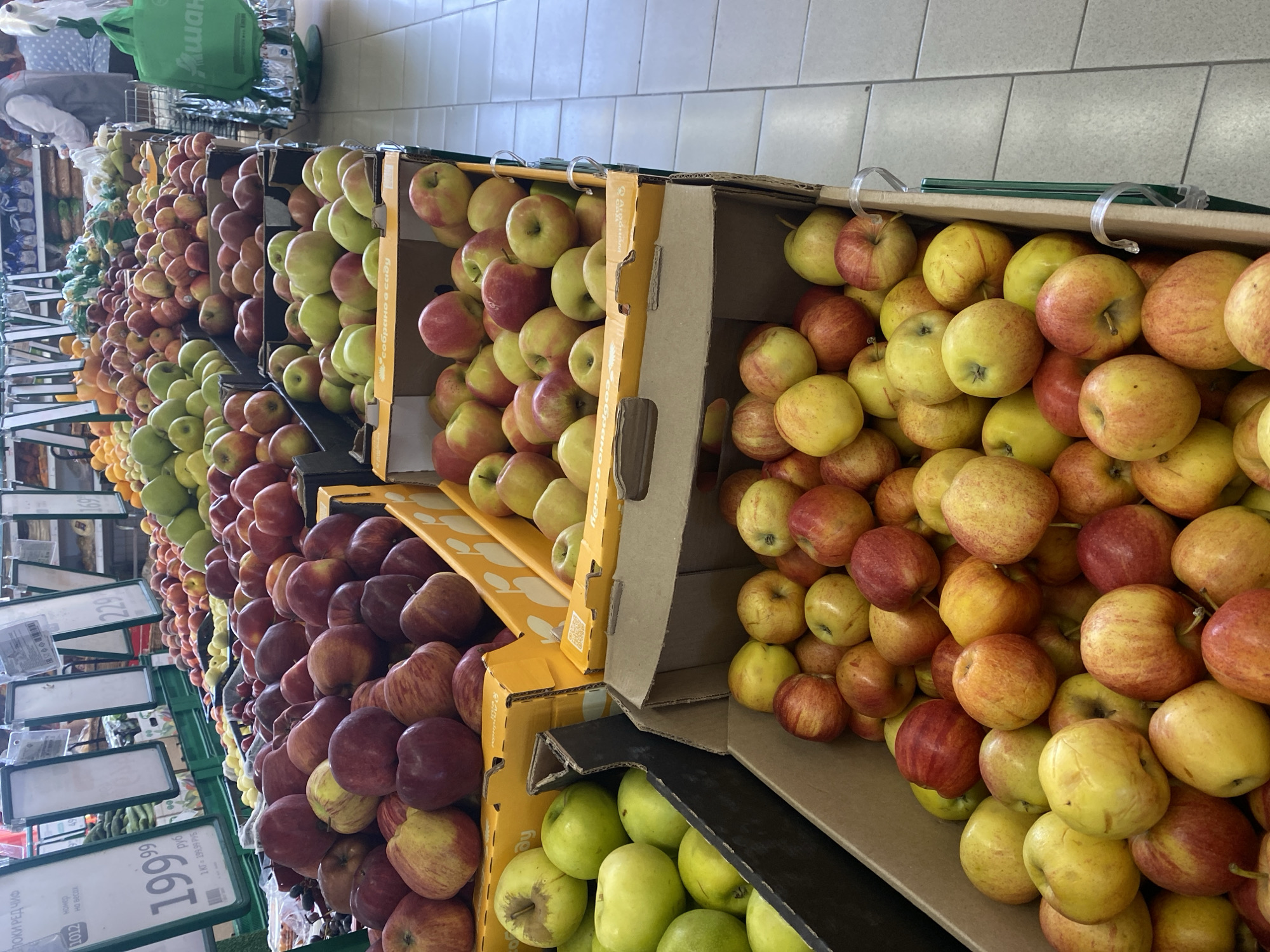 This is a medium shot of a produce section in a Moscow, Russia supermarket. In the foreground, several cardboard boxes are filled with apples. The boxes are stacked, with the top box in the center filled with red and yellow apples. To the left and right of this box, other boxes are also filled with apples of varying shades of red and yellow. Behind these boxes, more boxes of apples are visible, as well as some green apples in a black bin on the left. In the background, rows of more produce are visible, including what appear to be bananas and oranges. A person wearing a grey vest and a white shirt is visible in the far background, stocking shelves. A green shopping bag with Russian writing is hanging from a display rack on the far right. The flooring appears to be light-colored tile.