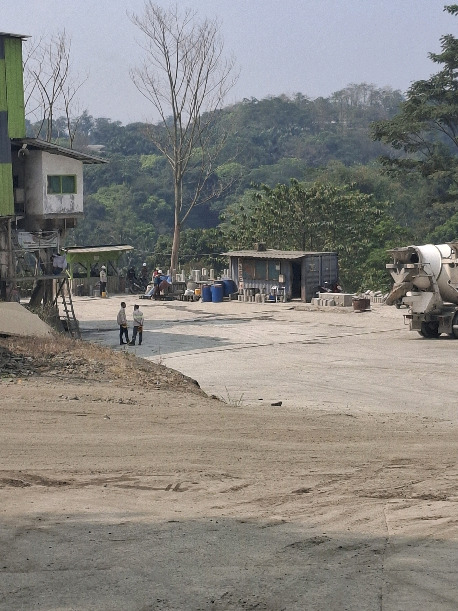 The image depicts an outdoor industrial site in Cijangkar, Indonesia. In the foreground, a dusty, uneven ground covers the majority of the frame, with some patchy vegetation and debris visible along the lower edge. In the middle ground, there is a paved area with several structures and vehicles. To the left, a multi-story building with green and black siding stands, featuring a small, elevated room with a green-framed window. A ladder is attached to the side of this structure. Nearby, a small, green-roofed stall is manned by a person in a white hard hat. Several other individuals, some in hard hats, are present, with one standing near a motorcycle and others near stacks of concrete cylinders and blue barrels. A cement mixer truck is positioned on the right side of the paved area, facing away from the viewer. In the background, a dense, green forest covers rolling hills, with a few bare trees protruding above the foliage. The sky is overcast and light.