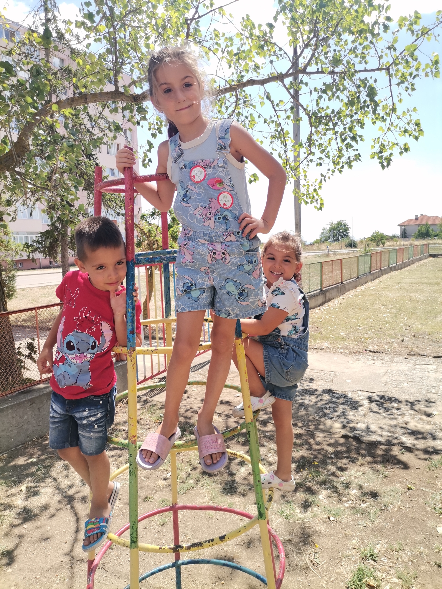 Three children are on a playground structure in Balchik, Bulgaria, on a sunny day. The tallest girl, standing in the middle, wears a light blue denim overall with a Stitch print and a white undershirt. She has blonde hair and is looking at the camera with a smile. To her left, a boy in a red t-shirt featuring Stitch and denim shorts is leaning against the structure, also smiling. To the right of the tallest girl, a younger girl in a white t-shirt with a Stitch print and blue denim overalls is smiling and looking at the camera while holding onto the structure. The playground has colorful metal bars. In the background, there are buildings, trees, and a fence.