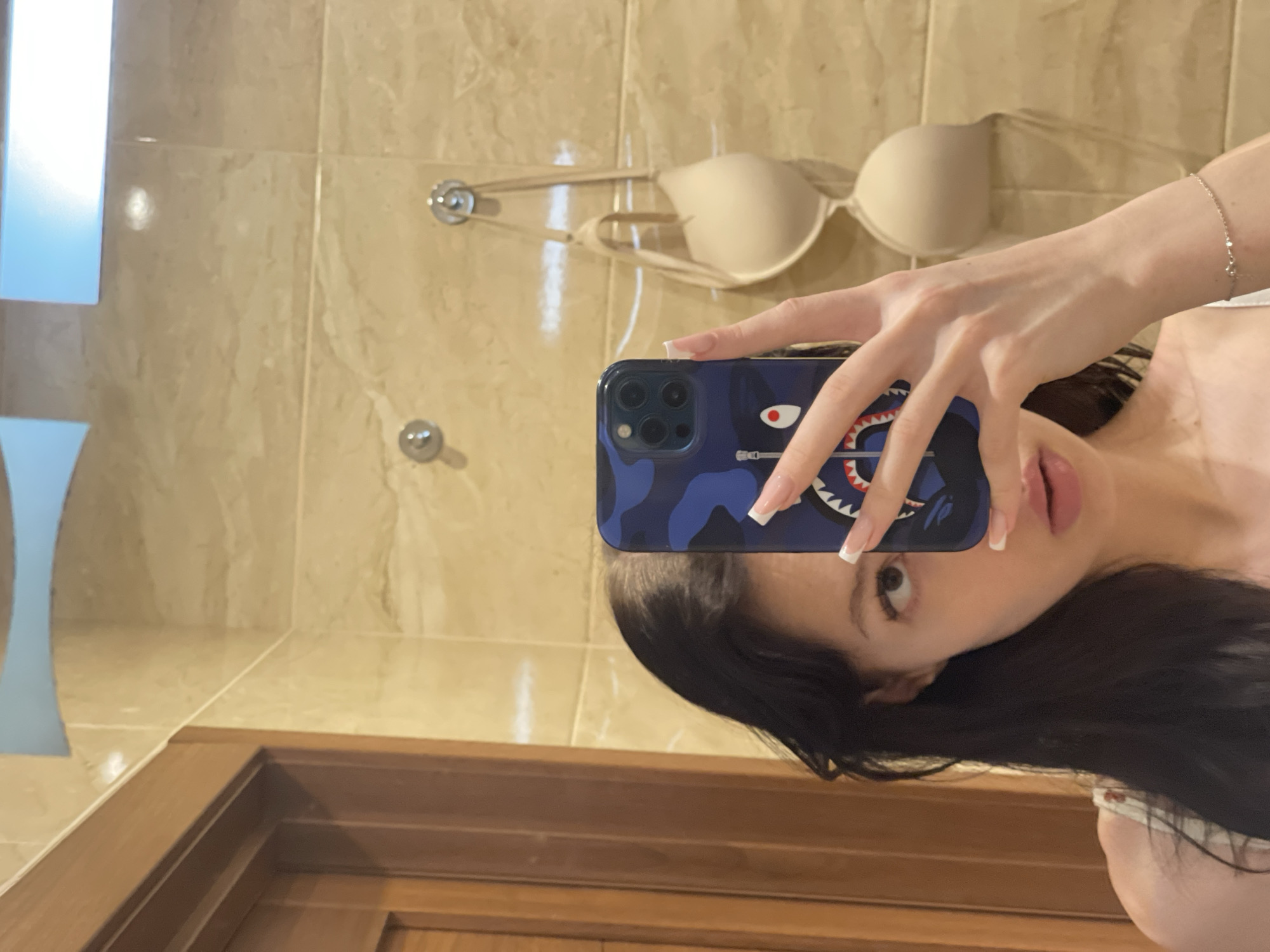 A young woman with dark hair is taking a mirror selfie in what appears to be a bathroom. She is holding a blue phone case with a shark graphic and is wearing a white top with a floral pattern. A beige bra is hanging on a hook on the wall behind her, which is tiled with marble-patterned tiles. To the left, a wooden door is visible. The lighting suggests an indoor setting, possibly a hotel room or a residential bathroom. The overall scene is casual and personal. There is no specific information in the image to indicate the location is in Khimki, Russia.
