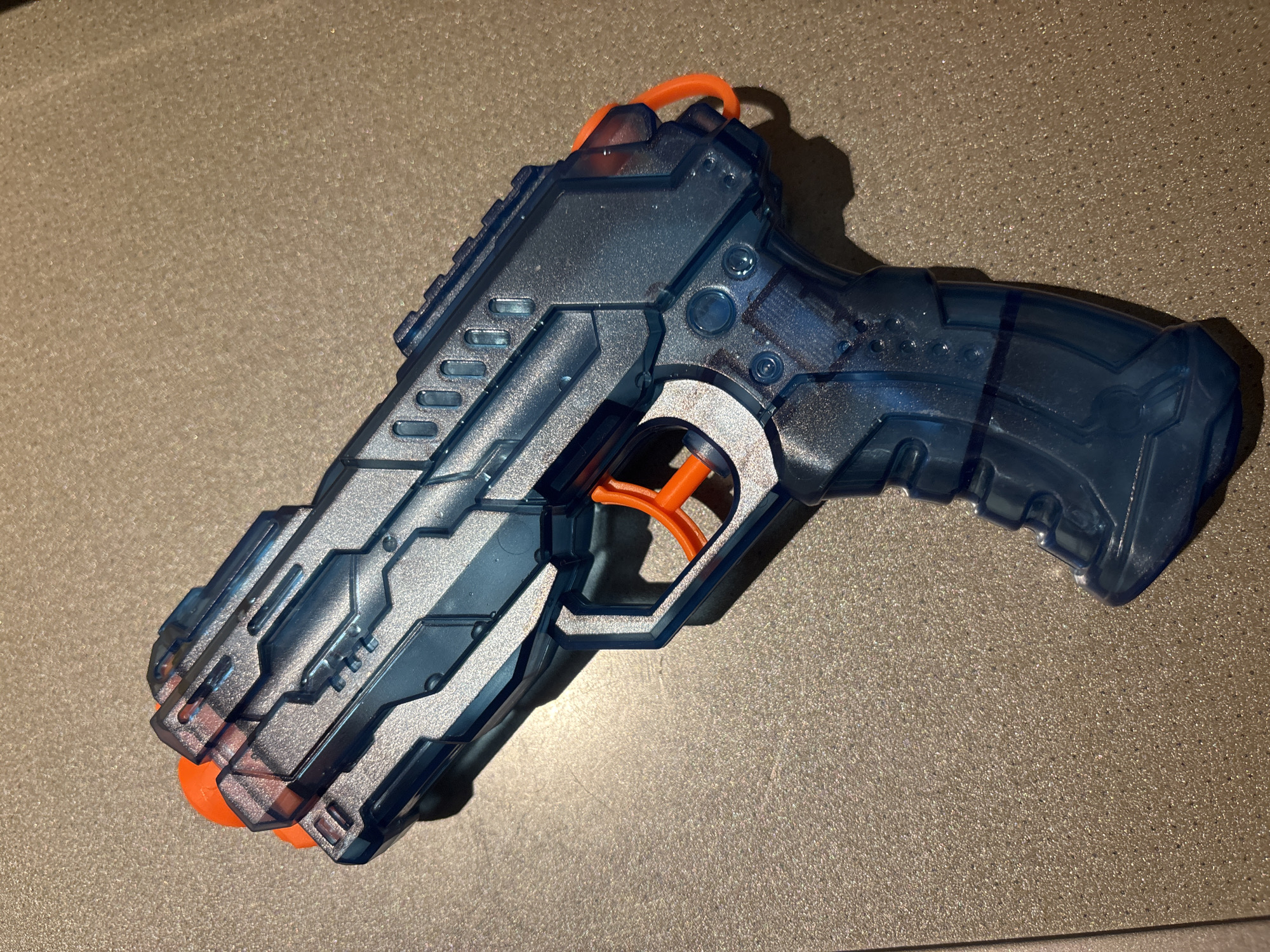 A blue and silver toy water gun with orange accents rests on a textured, speckled surface, possibly a countertop or floor. The toy is oriented diagonally from the upper left to the lower right of the frame. The gun features a transparent blue body with metallic silver detailing and has an orange trigger and barrel end. The background surface is uniformly lit, casting a soft shadow to the lower right of the object. The scene is indoors. No persons are visible. The location is not specified.