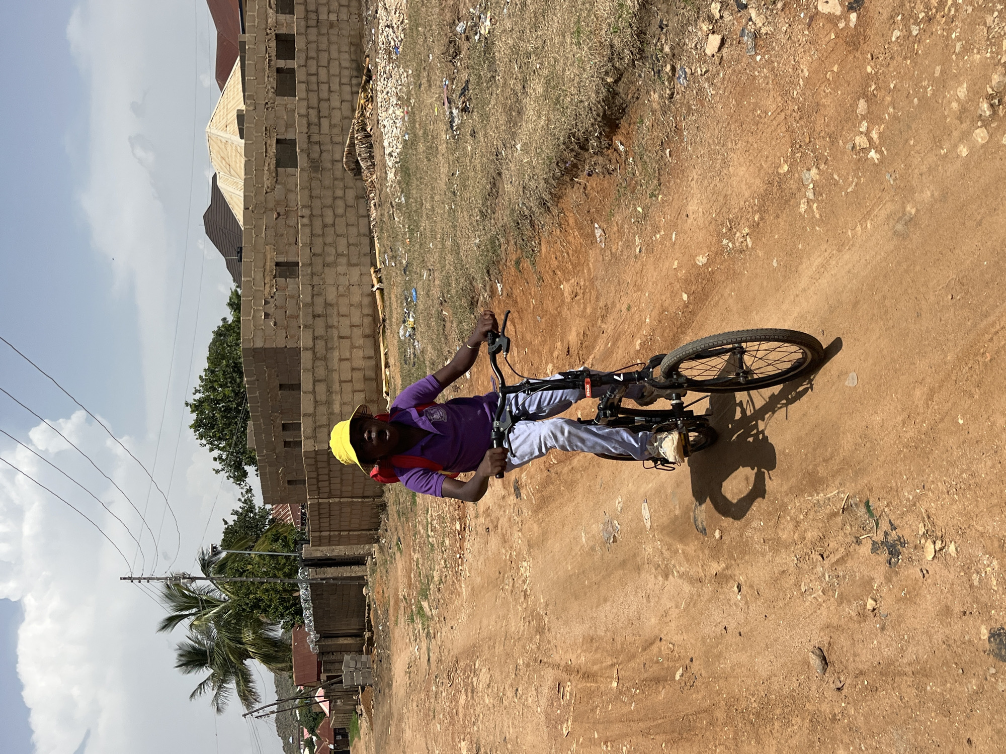 A young person wearing a yellow hat, a purple shirt, and light-colored pants rides a bicycle on a dirt road. The person is holding the handlebars and appears to be looking forward. A red backpack is slung over their shoulders. The background shows a dusty, unpaved road with scattered rocks and debris. In the distance, there are unfinished brick buildings, some with visible rebar, suggesting construction. There are also some trees and power lines visible. The sky is partly cloudy, indicating it is likely daytime. The overall impression is a scene in a developing area, possibly a rural or semi-urban environment. There is no visible text in the image.