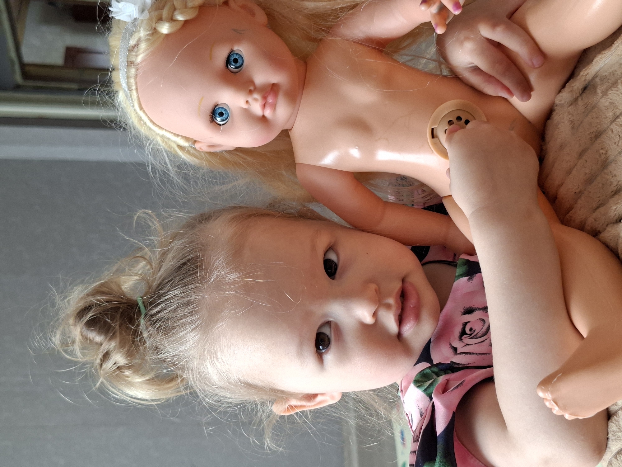 A close-up, indoor shot depicts a toddler with blonde hair, styled in a bun secured with a green hair tie, wearing a pink floral-patterned shirt. The toddler lies next to a blonde doll with blue eyes and a plastic body. The toddler's hands are positioned on the doll's stomach, near a circular, grid-like opening on the doll's abdomen. The background shows a beige, textured surface and a partially visible framed object. The location and city are unknown.