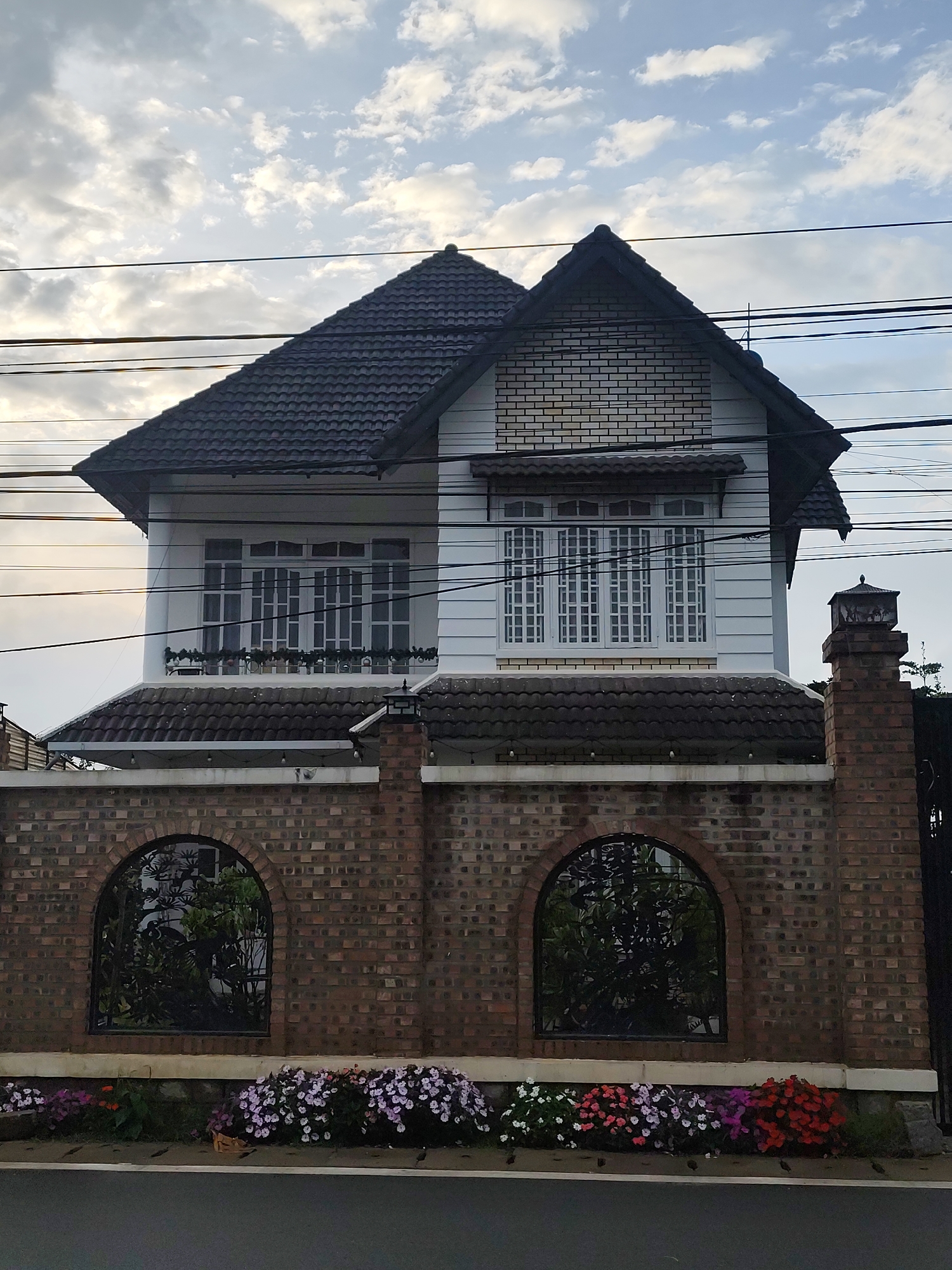 The image depicts a two-story house in Lâm Viên - Đà Lạt, Vietnam, viewed from across a street. The house has a dark, tiled roof and white siding. The upper floor features several windows with white frames and a decorative railing with potted plants. The lower portion of the house is partially visible above a brick wall. The wall is constructed from reddish-brown bricks and features two arched windows with ornate metal grills, through which greenery is visible. A brick pillar with a decorative lantern stands to the right of the house. A colorful bed of flowers, including pink, white, purple, and red blossoms, lines the street in front of the brick wall. Overhead, power lines crisscross against a cloudy sky. The scene appears to be taken during the daytime, with soft, diffused lighting. There are no people visible in the image.