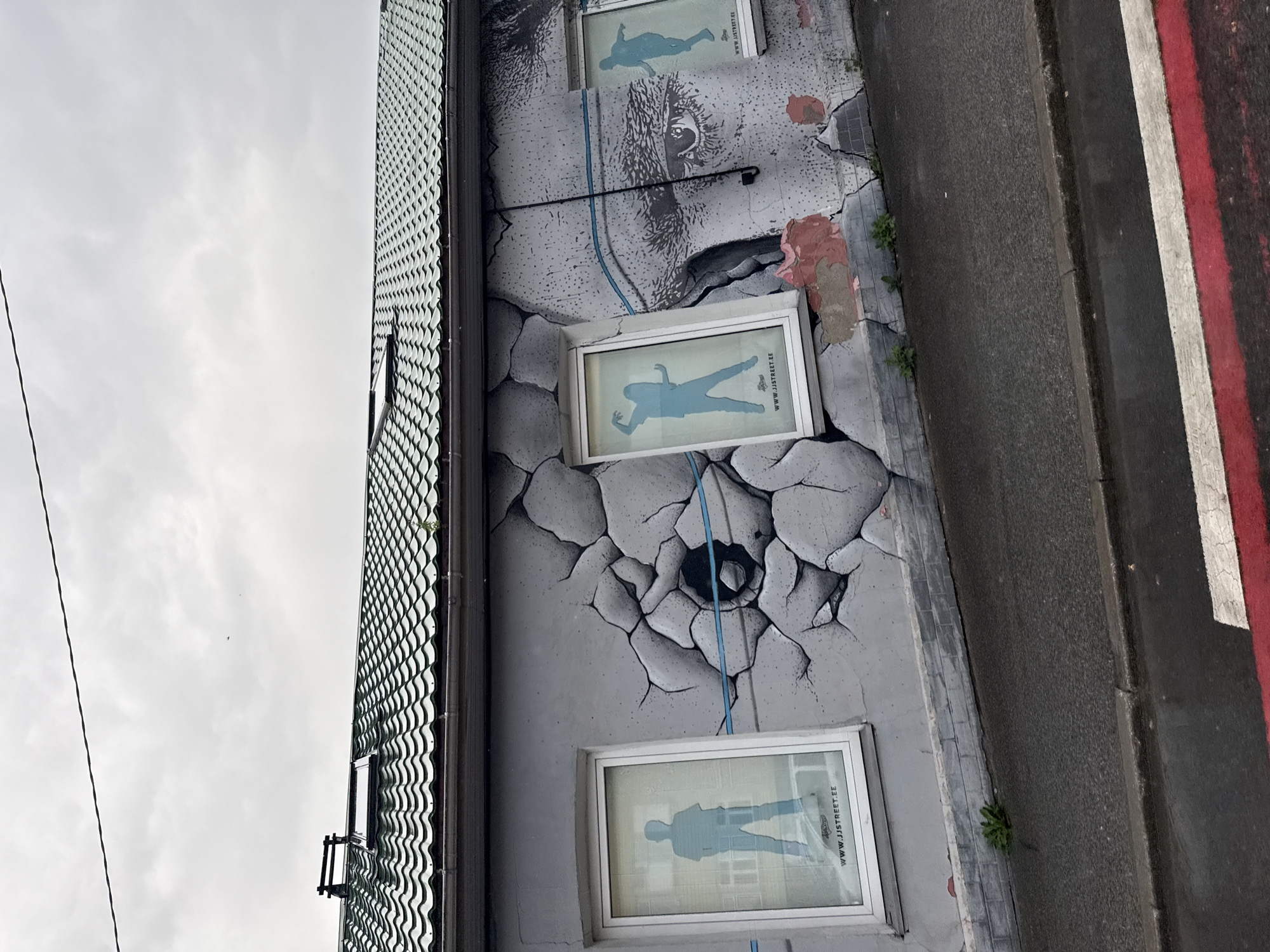 This image depicts a building facade in Tallinn, Estonia, adorned with a large, striking mural. The mural features a hyperrealistic, cracked concrete effect, giving the impression that the wall is breaking apart to reveal an inner structure. Within these cracks, two windows are integrated, each displaying a silhouette of a person in a dynamic pose. These silhouettes are rendered in a bright blue. The larger window on the left shows a person standing with hands at their sides, while the window on the right shows a person in a dance-like posture. Below each silhouette, the website "WWW.JJSTREET.EE" is visible, suggesting this is an advertisement or a promotion for a street dance school or event.
The mural also extends to cover a significant portion of the wall between the windows, incorporating a large, detailed depiction of an eye, with textured shading and apparent distress or age lines. A thin blue line runs horizontally across the face and the cracked concrete sections, adding a graphic element. Some areas of the mural show patches of pink and red paint, as if simulating peeling paint or plaster.
The building itself has a green, tiled roof with a skylight and visible guttering. The facade is painted a light grey, providing a backdrop for the mural. Below the building, a wet asphalt sidewalk runs alongside the street, marked with red and white crossing lines, indicating a pedestrian crossing. The sky is overcast, with a few faint clouds. The overall impression is one of urban art transforming a mundane building into a visually engaging spectacle.