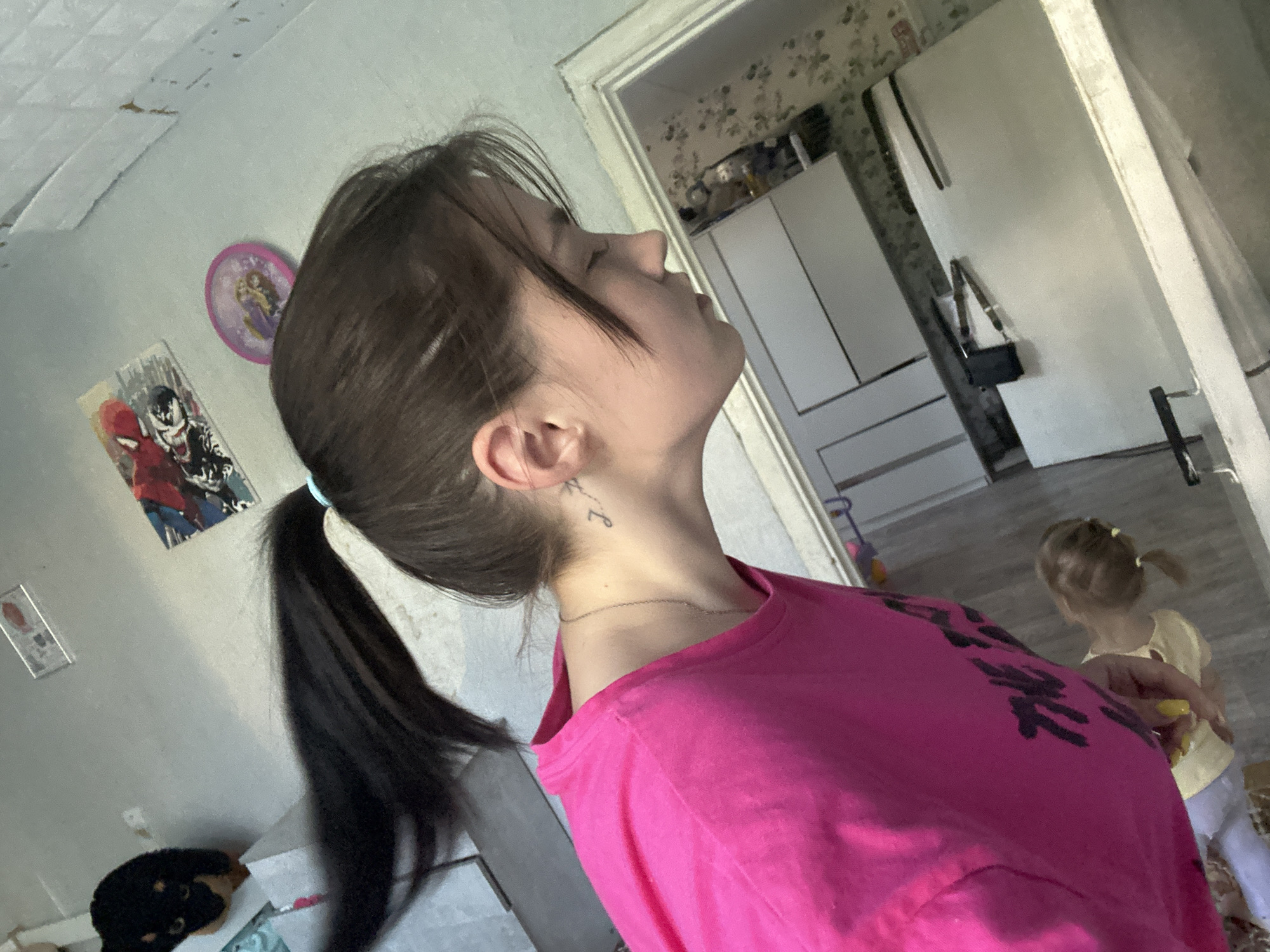 The image is a medium shot, taken indoors, of a young woman with dark hair in a ponytail, wearing a bright pink t-shirt. She is facing away from the camera, with her head tilted back and eyes closed. A small tattoo is visible on the back of her neck. In the background, a young child with blonde hair in a ponytail is visible, wearing a yellow shirt and light-colored pants. The environment appears to be a bedroom or living area, with wallpaper visible on the walls and furniture in the background. There are posters or pictures on the wall. The lighting suggests it is daytime. The image is from Chaikovsky, Russia.