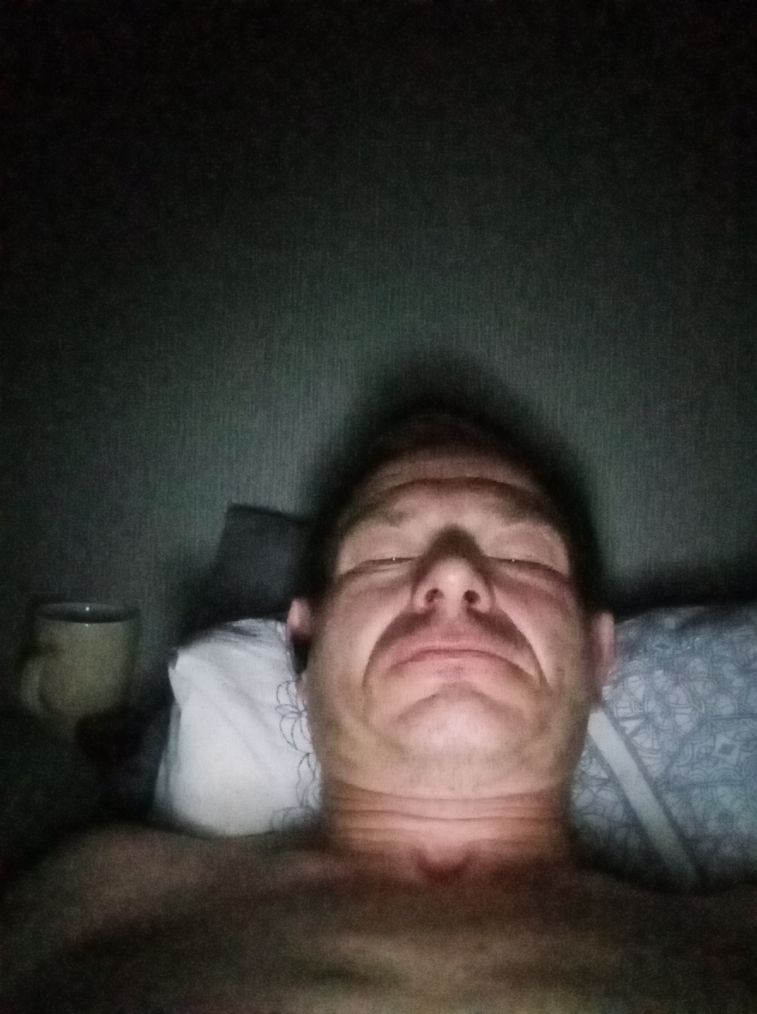 The image is a close-up, low-light, selfie-style photograph of a man lying down. The man is positioned centrally in the frame, with his head tilted slightly back, and his eyes are closed. His facial expression appears contorted, possibly due to the camera angle or a relaxed, drowsy state. A mug is visible to the left of the man's head. The background consists of a textured wall and what appears to be bedding, including a pillow with a patterned design. The overall lighting is dim, with shadows obscuring some details. The image does not contain any information that would allow for its location to be specified within the city of Tapa, Estonia, or any other geographical location.
