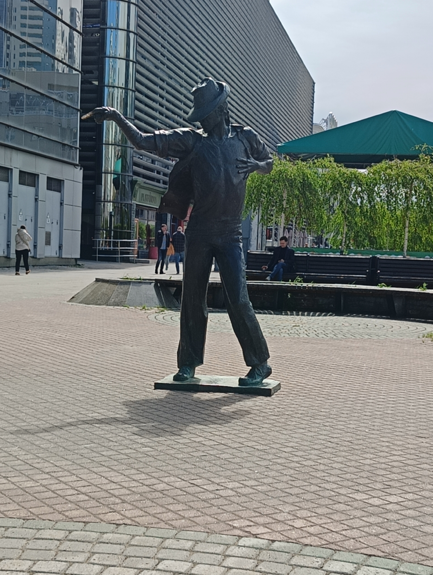 A full shot captures a bronze statue of a man wearing a hat, shirt, jacket, and trousers. His left arm is extended pointing towards a modern glass building, while his right hand rests on his chest. The statue stands on a square bronze base on a brick paved square. Behind the statue is a seating area with benches and trees, and a green roof is visible in the background. People are seen walking and sitting on the benches. The statue, located in Yekaterinburg, Russia, commemorates Michael Jackson.