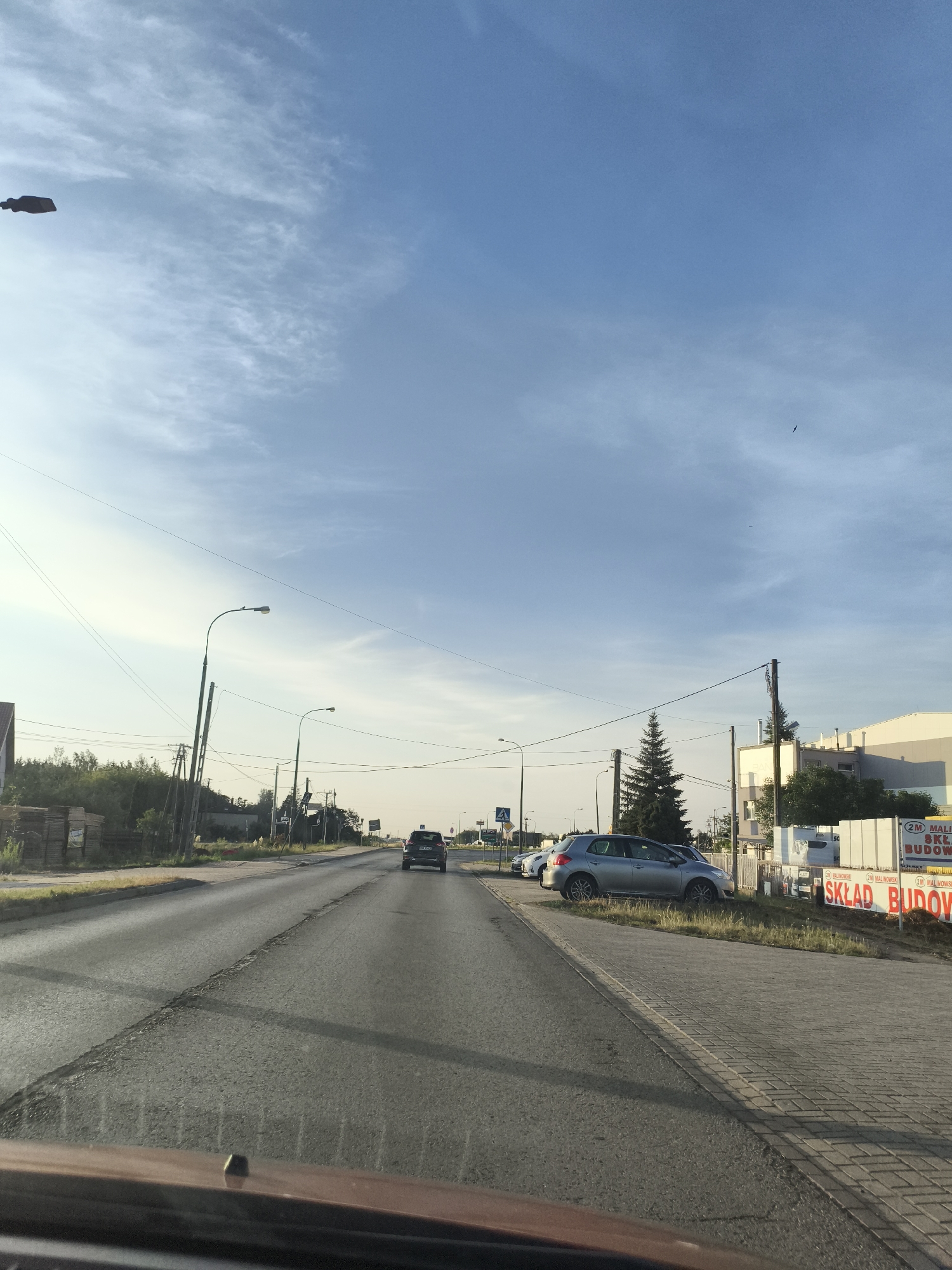 The image captures a daytime scene on a two-lane asphalt road in Czosnów, Poland. The road appears to be an arterial route with parked cars and buildings lining the right side. The sky is a clear blue with scattered wispy clouds.
On the left side of the road, there are stacks of what appear to be building materials, possibly lumber or palettes. Further down the road, there are trees and some low-rise buildings. Several utility poles with electrical wires are visible along the roadside.
On the right side of the road, there is a sidewalk paved with interlocking bricks. Parked cars are visible along this side, including a silver hatchback and a dark-colored SUV. A large, white building with some signage is also present. One sign, in red lettering, reads "SKŁAD BUDOWY" which translates to "BUILDING SUPPLY STORE," and "MALINOWSKI". There is a tall, dark green evergreen tree in front of this building.
The perspective suggests the image was taken from inside a vehicle, looking forward. The road surface shows some wear and tear, with cracks visible in the asphalt. The overall impression is a commercial or semi-industrial area on the outskirts of a town.