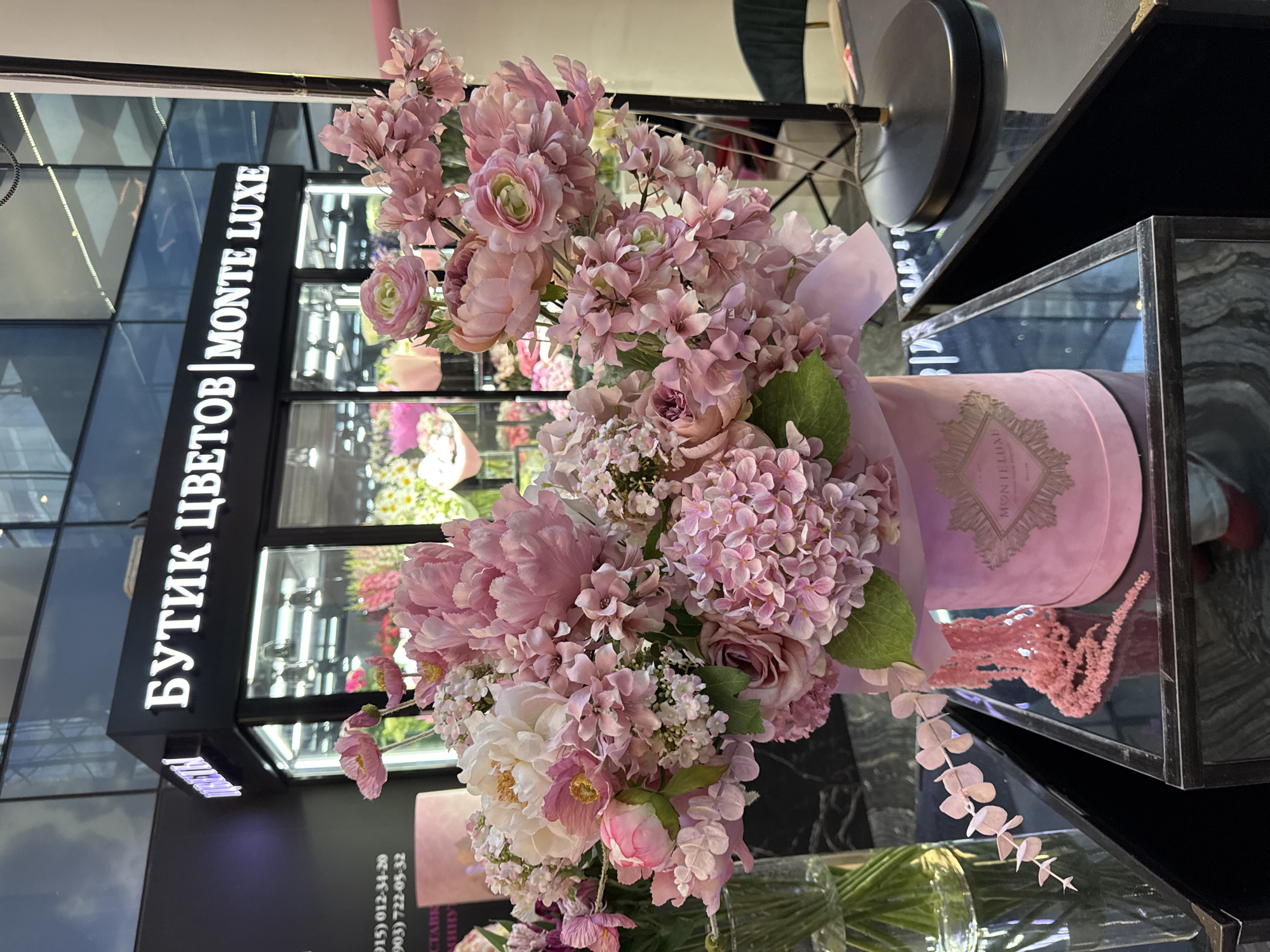 The image captures an elegant display of flowers within an indoor setting, likely a high-end flower boutique. The primary subject is a large, opulent bouquet, predominantly composed of pink, white, and light purple blossoms with lush green foliage. Various flower types are visible, including full-petaled varieties resembling peonies and ranunculus, clusters of hydrangeas, and other smaller, delicate blooms.
The bouquet is presented in or beside a luxurious, round, pink velvet hatbox, which features a golden emblem or logo on its side, adding a touch of sophistication. Adjacent to the hatbox, a decorative element of pink feathery or dried foliage can be seen, partially reflected on a sleek, black, marble-patterned surface. In the lower part of the frame, a glass vase holding green stems is visible, along with a delicate branch featuring light pink leaves or petals.
The background features a tall, black sign with vertically illuminated white text that reads "БУТИК ЦВЕТОВ | MONTE LUXE," which translates from Russian to "Flower Boutique | MONTE LUXE." Below this sign, on a dark surface, two phone numbers are listed: (915) 012-34-20 and (903) 722-05-32. The setting includes modern display cases with reflective glass and dark frames, creating a contemporary and polished environment. Artificial lighting illuminates the scene, highlighting the vibrant colors of the flowers. No people are present, and the image solely focuses on the detailed presentation of the floral arrangement and the boutique's branding. The overall atmosphere conveys luxury and meticulous design.