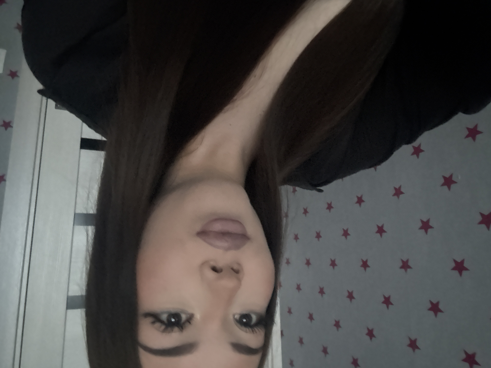 The image shows a close-up, selfie-style photograph of a young woman with dark hair and makeup. She is wearing a dark top. The background consists of a wall with a pattern of pink stars on a light gray surface, and to her right, a portion of a white door or wall with vertical lines. The lighting is somewhat dim. The image does not contain any information that would allow for its location to be identified as Naberezhnye Chelny, Russia.