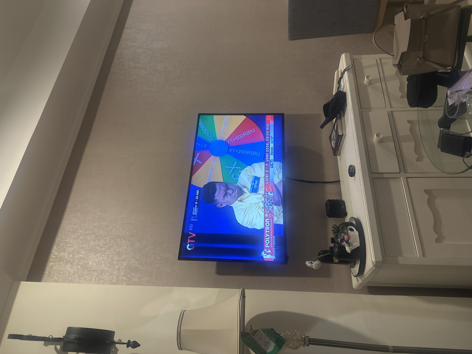 The image shows a television mounted on a wall in a room, displaying a program with a wheel of fortune graphic and a person identified as "OPAN." Text on the screen includes "GTV HD AFC U17 ASIAN CUP 6-22 MEI POLYTRON INDONESIA OPEN 26 TRANSVISION," along with prize amounts on the wheel, such as "X1+500RIBU" and "X1+250RIBU." The program also indicates a date and time: "LIVE! 2-7 JUNI 2026, 09:00 WIB RCTI iNews RCTI."
Next to the television is a white dresser with several drawers and items placed on top, including a black cap, a picture frame, and a vase with flowers. On top of the dresser are bags and electronics. A lamp with a white lampshade is visible in the lower part of the image.
Based on the text "POLYTRON INDONESIA OPEN 26" and the presence of RCTI (a TV station from Indonesia), it's plausible that the location is within Indonesia. However, the specificity to confirm it is Central Jakarta is not evident within the image itself.