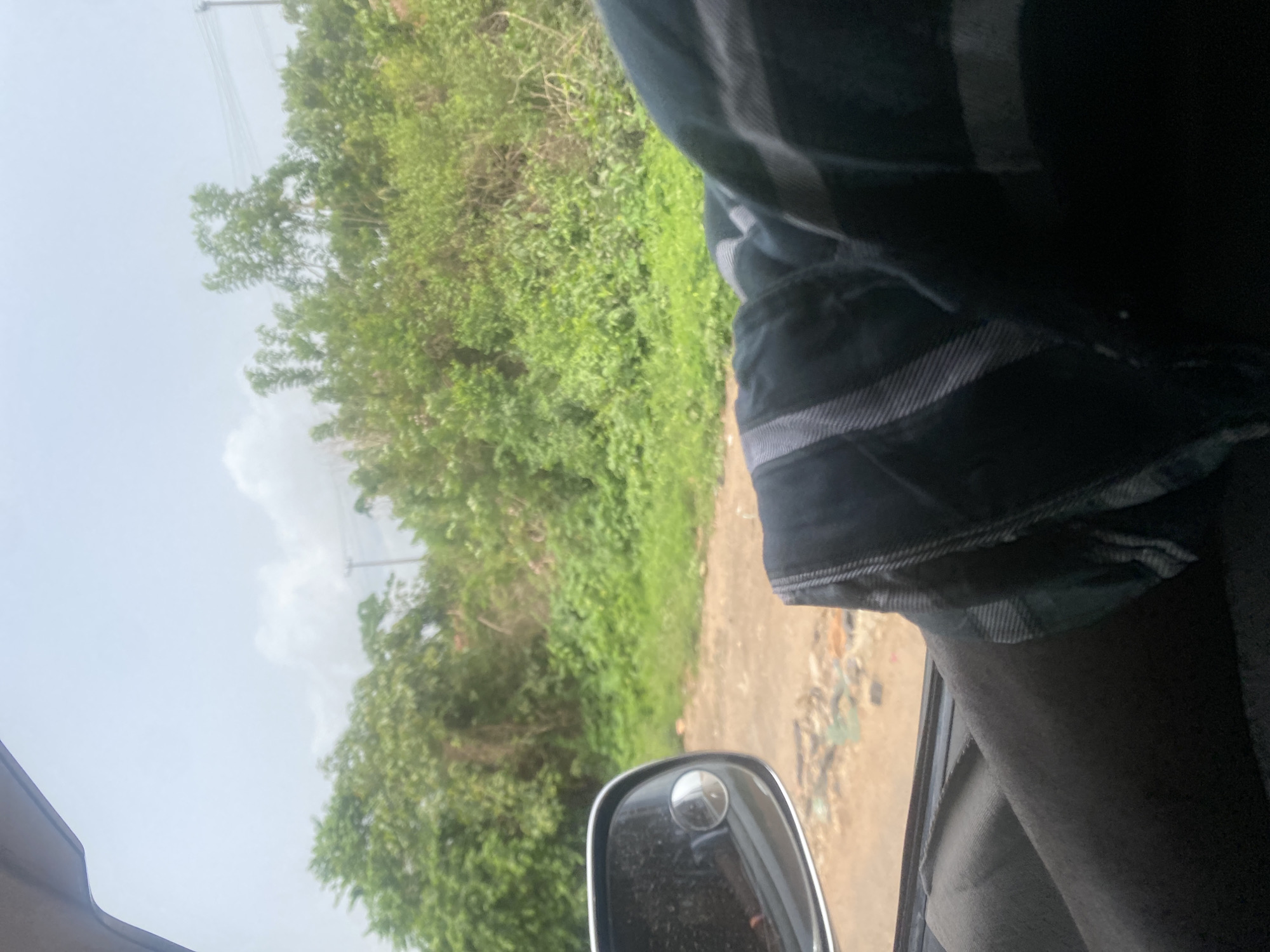 The view is from inside a moving vehicle, likely a car, driving on a road in a lush, green area. The side mirror of the vehicle is visible. The road appears unpaved and there's possible debris or litter on the roadside. Outside, dense foliage of trees and bushes lines the road. In the distance, utility poles can be seen. The sky is overcast. There's no other contextual information to identify the precise location in Osu, Nigeria. No people are distinctly visible, apart from the arm with a plaid/striped pattern shirt.
