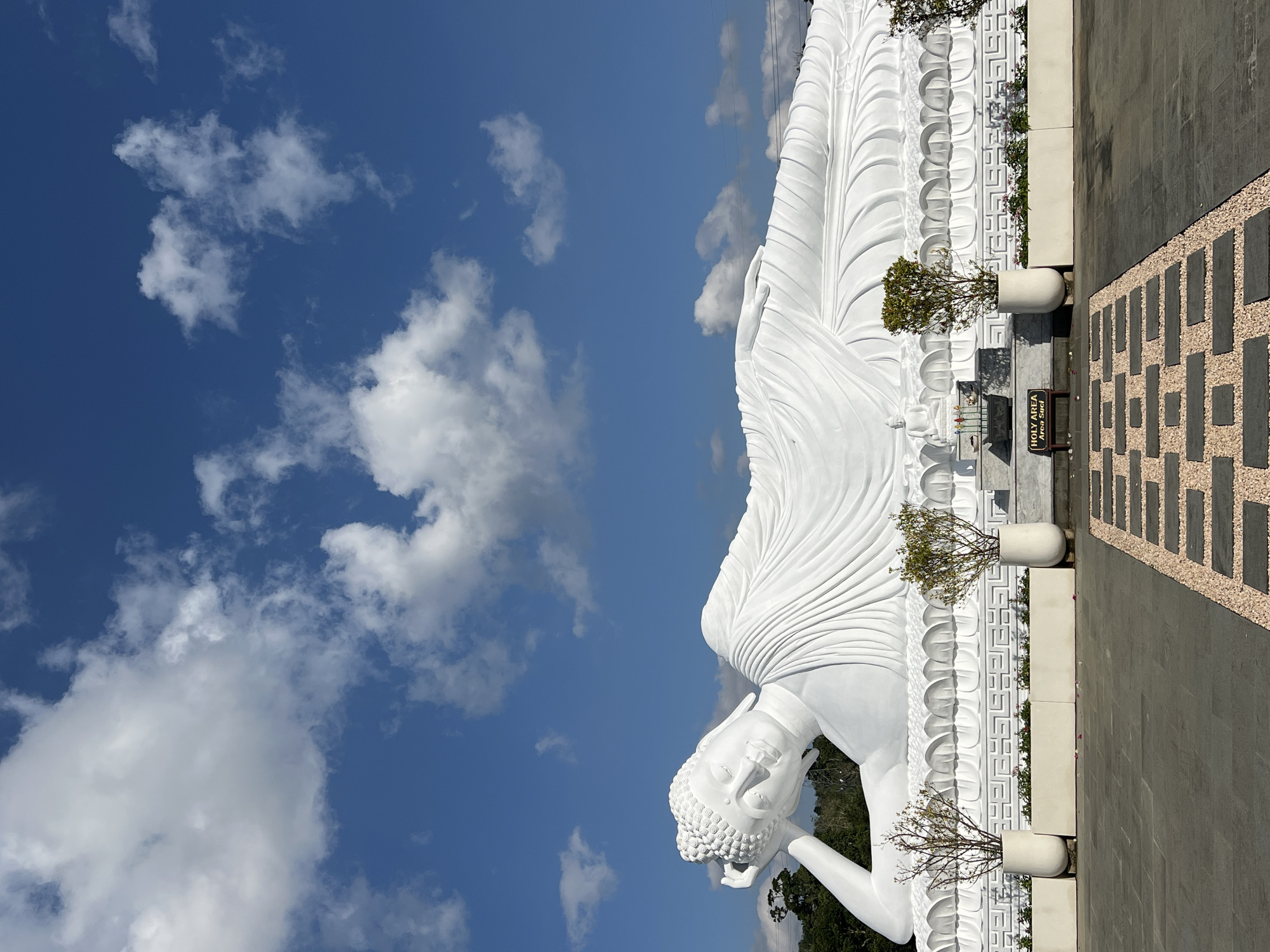 The image shows a large, white, reclining Buddha statue against a bright blue sky with scattered white clouds. The statue is a prominent feature, taking up a significant portion of the frame. The Buddha is depicted in a serene posture, with its head resting on its arm and its body elongated. The statue is highly detailed, with intricate folds in its robes and a sculpted facial expression.
In front of the statue, there is a stone base with decorative carvings. Two tall, white planters containing small trees are placed on either side of a low, rectangular planter filled with small pink flowers. Between the planters, there is a small altar with offerings. A sign in front of the altar reads "HOLY AREA Area Suci".
The foreground of the image features a paved walkway with a patterned arrangement of dark gray stones interspersed with light brown gravel. The ground is paved with dark gray stone tiles.
The background shows a line of lush green trees, indicating an outdoor setting. There are also some power lines visible in the distance.
This statue is located at the Pujayama Brahma Vihara in Pupuan, Bali, Indonesia. The area appears to be a religious or spiritual site, given the presence of the statue, altar, and signage. The overall impression is one of peace and tranquility.