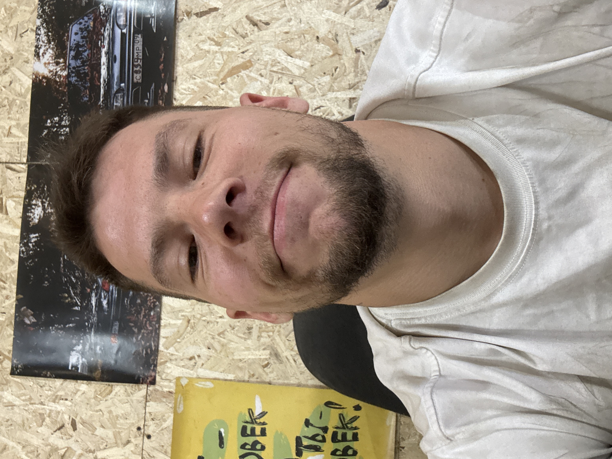 A close-up, rotated image features a fair-skinned male with short brown hair, a beard, and a slight smile. He is wearing a white t-shirt. Behind him, affixed to a light-colored particleboard wall, are two posters/images and a yellow object. One poster depicts a black vehicle, possibly a car, with a license plate visible reading "C 859 MP 09". The other poster is mostly obscured but appears to be a yellow rectangular object with green and black markings on it.
The location in the city of Chaikovsky, Russia is not determinable from this image.