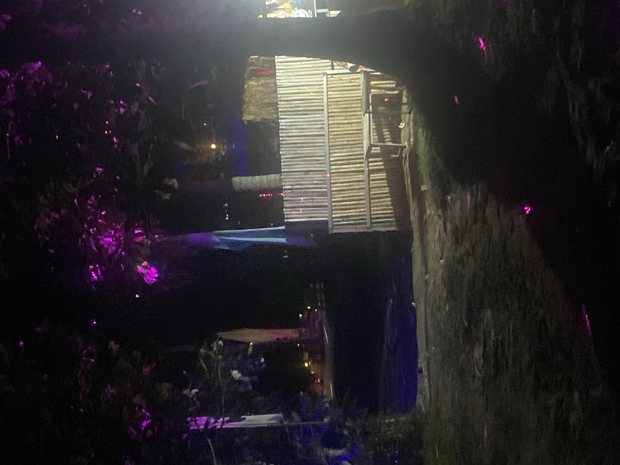 The image depicts a dimly lit scene at night in Ban Haad Rin, Thailand. A structure resembling a small building or platform made of bamboo-like material is visible above what appears to be a body of water. The water reflects some of the light, creating a mirrored effect of the structure. The foreground and the sides of the image are shrouded in darkness, with trees or dense vegetation, some illuminated by purple lights. A walkway or staircase is attached to the structure. The environment suggests a natural or semi-natural setting, possibly a tourist area given the lighting and structure.