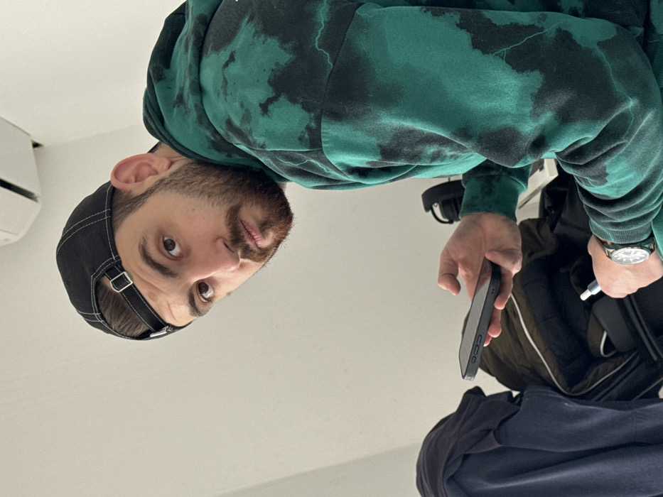 An indoor scene features a male subject. He is wearing a black baseball cap reversed on his head and a green and black tie-dye hooded sweatshirt. A silver-toned wristwatch is on his left wrist. The subject is looking directly forward, holding a dark-cased smartphone horizontally in his right hand and a silver-tipped vape pen in his left hand. The background is a plain white wall. An off-white wall-mounted air conditioning unit is visible in the upper right. In the lower left background, dark fabric items, including a dark green jacket, are present. The precise location within the city of Varna, Bulgaria, is not discernible from the visual evidence provided.