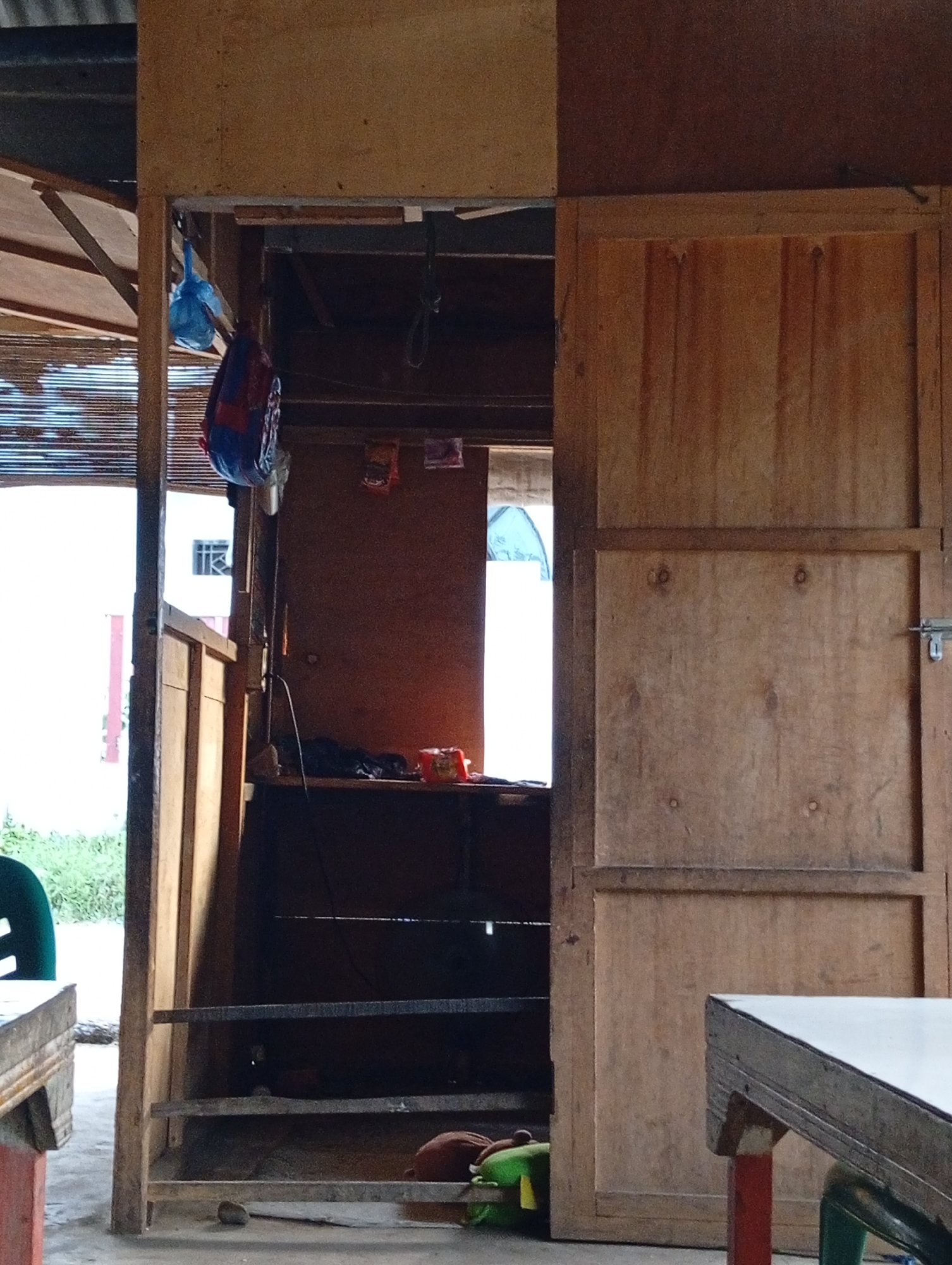 The image depicts an outdoor setting, likely a stall or small shop, constructed with unfinished wood. In the foreground, a portion of a table with a white surface is visible on the right. To the left, another table with a similar white surface is partially visible. The main structure features an open doorway leading to an interior space. Inside, shelves are stacked with various items, including what appear to be bags and food packaging. Two blue plastic bags hang from the ceiling of the interior. A closed wooden door with a latch is visible on the right side of the structure. The background reveals a white building and greenery, suggesting an urban or semi-urban environment. There are no people visible in the frame. The overall scene is unkempt, with visible dust and a general air of casual use. This scene could be in Lhokseumawe, Indonesia, given the visual cues of simple, open-air structures common in such environments.