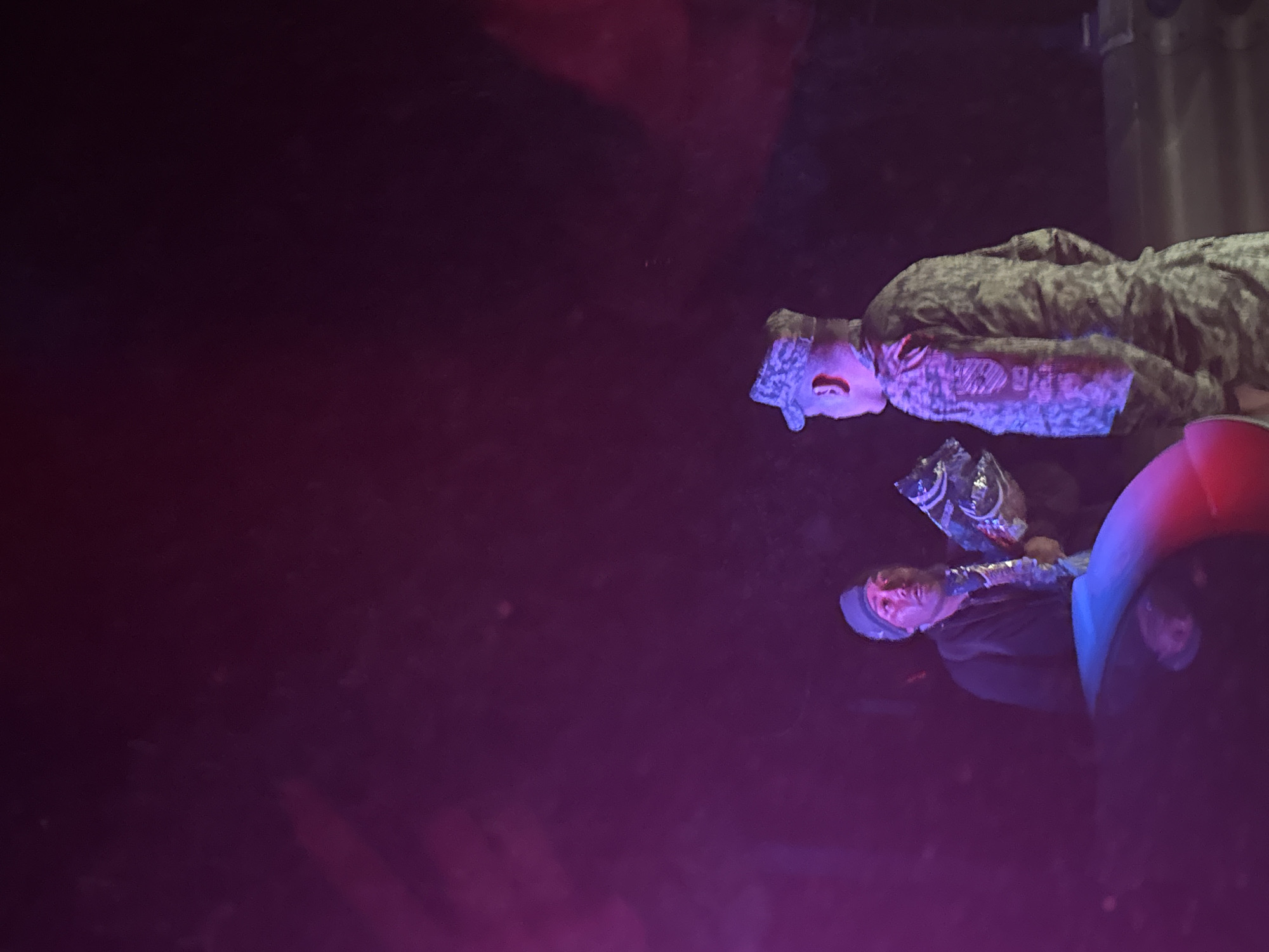 The image displays three individuals in a dimly lit indoor setting, illuminated by vibrant purple and pink stage-like lighting. The overall atmosphere suggests an evening event or performance.
On the left side of the frame, a person is prominently visible, wearing a camouflage-patterned jacket and a matching cap. Their face is brightly lit by the colorful light, and their mouth appears slightly open, as if speaking or singing.
In the center, another individual wears a dark cap and a dark jacket. They are holding a crumpled, possibly reflective, plastic bag or object in their hands. A shiny, metallic-looking accessory is visible around their neck or chest. Their face is also highlighted by the strong purple and pink illumination.
A third person is partially seen on the right, with only their head and upper torso visible. They are wearing a garment that combines red and blue colors.
The background is largely dark and indistinct, with a tall, dark, cylindrical object faintly visible in the upper right, possibly a structural element or equipment within the venue. There is no discernible text anywhere in the image. The scene captures a moment from what appears to be a social or entertainment event in San Marcos Huixtoco, Mexico.