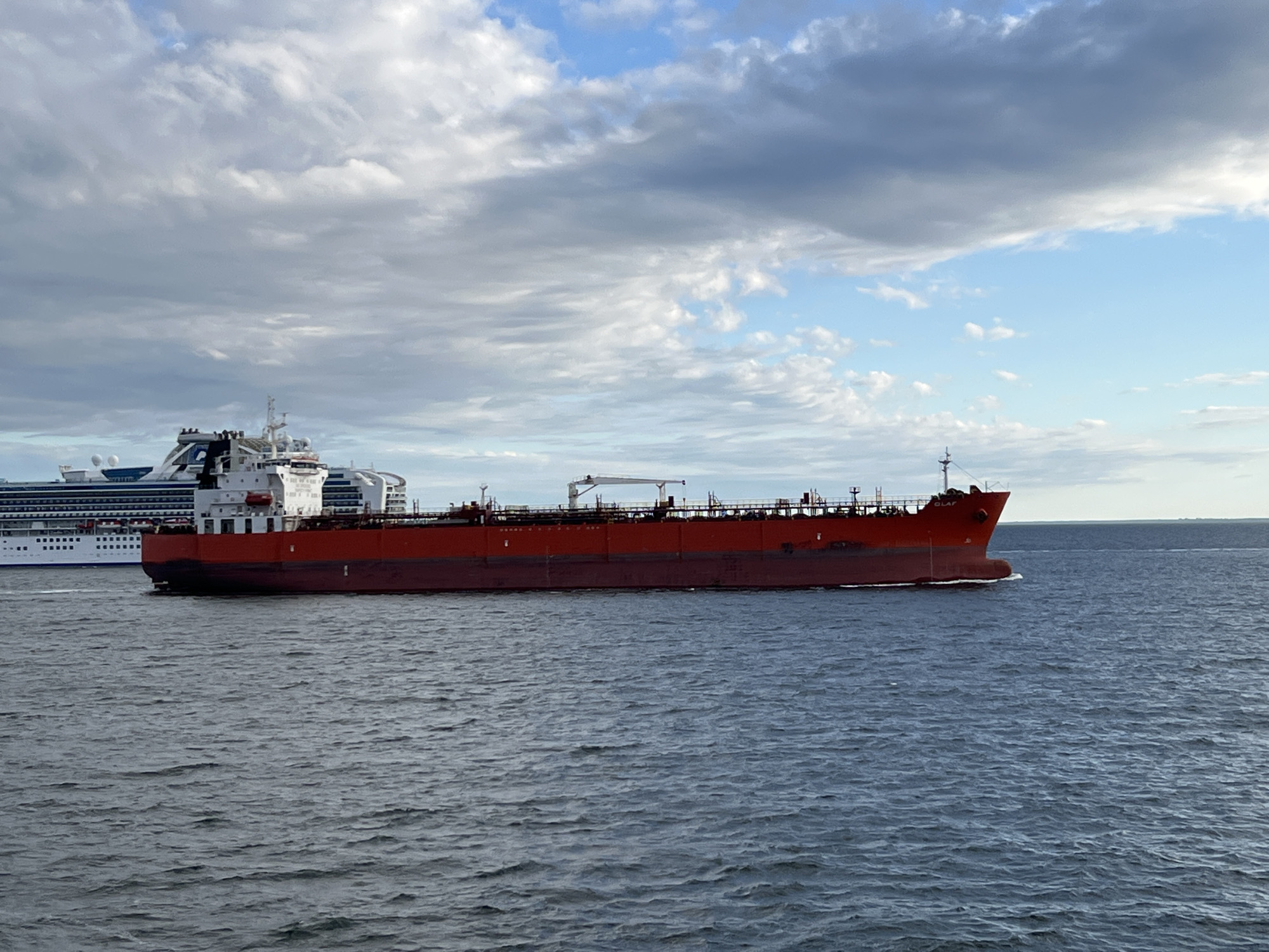 A large red tanker ship sails on a blue body of water under a cloudy sky. In the background, a white cruise ship is docked. The tanker ship is the primary focus of the image, with its hull extending horizontally across the frame. The ship has a white superstructure and a crane. The water is choppy, with small waves. The sky is partly cloudy, with patches of blue sky visible. The image is taken from a low angle, looking up at the ships. The scene is likely set in the port of Gdynia, Poland, given the presence of both tanker and cruise ships, which are common in the city's port.