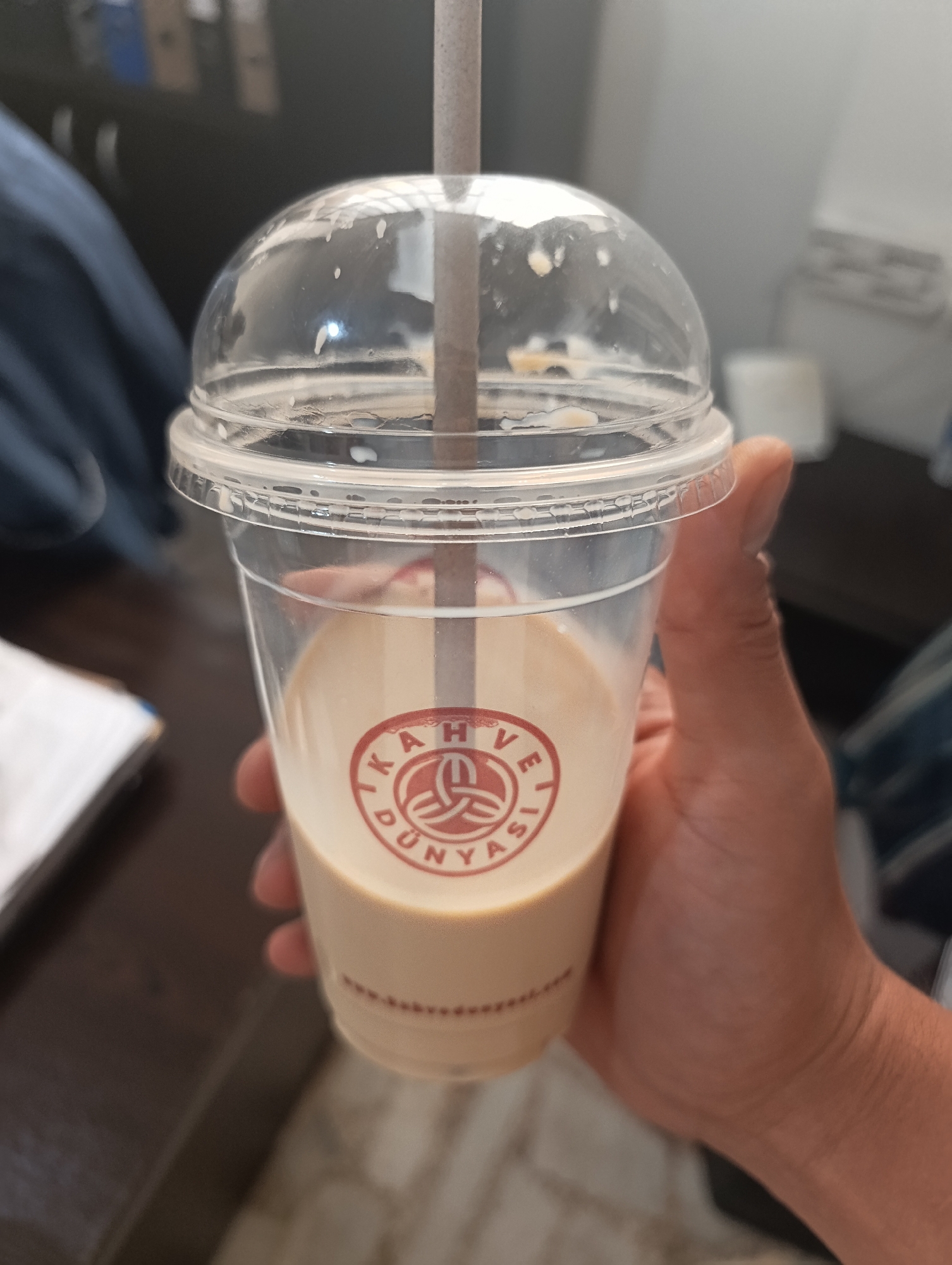 A person is holding a clear plastic cup with a domed lid and a straw. The cup contains a light brown beverage, possibly coffee or tea with milk. A red logo with text in Turkish, "KAHVE DÜNYASI", is visible on the cup. The background is blurred, showing a dark surface that could be a desk or table, and some office-like elements. The image suggests the person is in an indoor setting, possibly an office or a cafe. No specific location in Ümraniye, Turkey is discernible from the visual information.