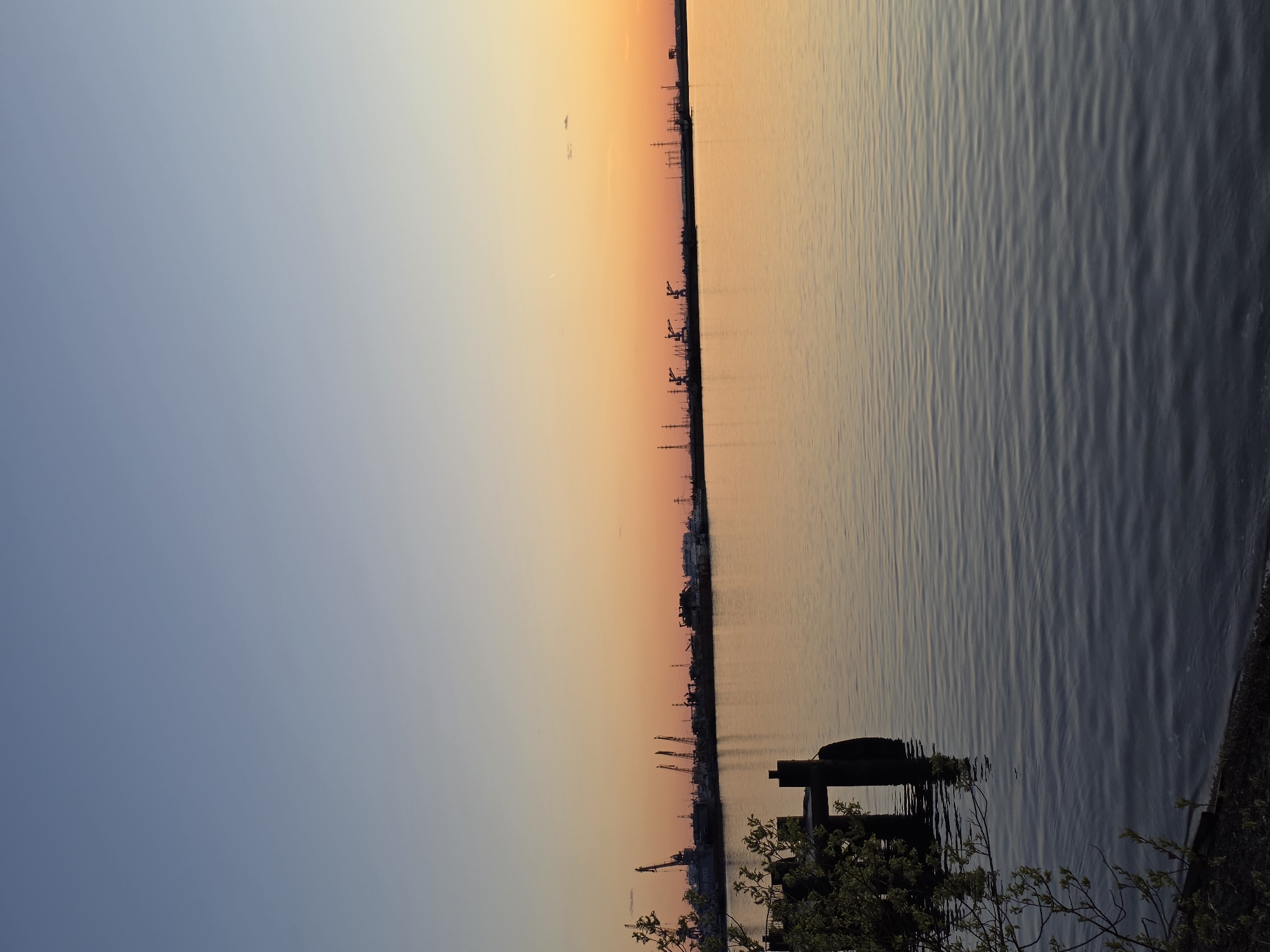 The image shows a body of water, likely a river or large lake, under a sunset or sunrise sky. The sky transitions from a deep blue on the left to orange and yellow hues on the right. A silhouette of a city skyline is visible on the horizon line, featuring cranes and industrial structures, indicating a port or industrial area. In the foreground, there is some vegetation and a wooden structure partially submerged in the water. The water is calm with ripples and reflections of the sky. Based on the prompt, the location might be the city of Chaikovsky, Russia.