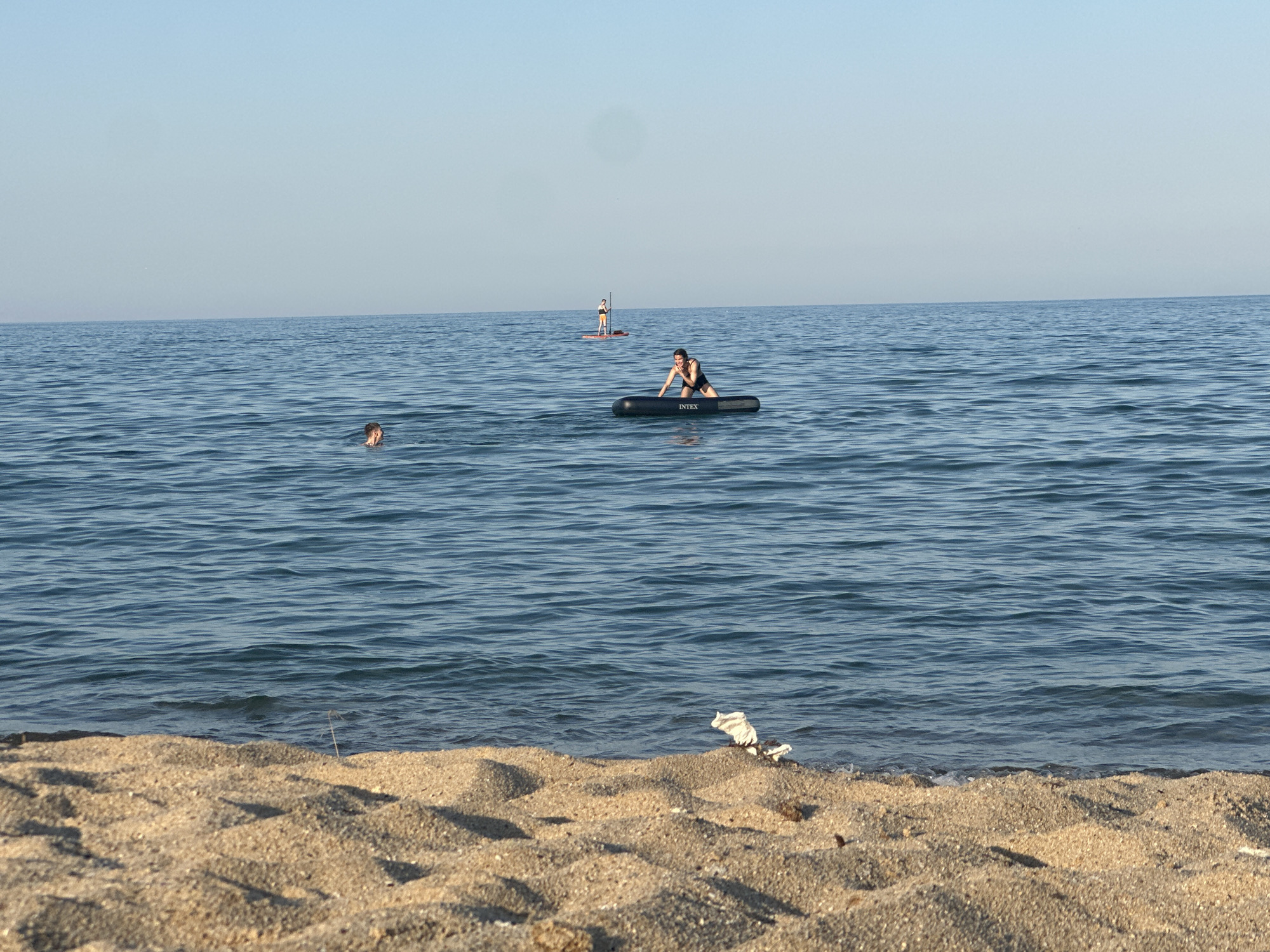The image depicts a serene scene at a beach with calm blue water. The foreground is dominated by a sandy shore, with a piece of white debris visible near the waterline. In the middle ground, several people are enjoying the water. One person is partially submerged in the ocean, with only their head visible. Another individual is seated on an inflatable black raft, which has the brand name "INTEX" clearly printed on it. In the background, two people are standing on paddleboards, further out in the water. The sky is clear and a pale blue, suggesting it is daytime, likely morning or late afternoon due to the soft lighting. The overall mood is peaceful and recreational, with individuals engaging in various water activities. The location appears to be a coastal area with a calm sea.
