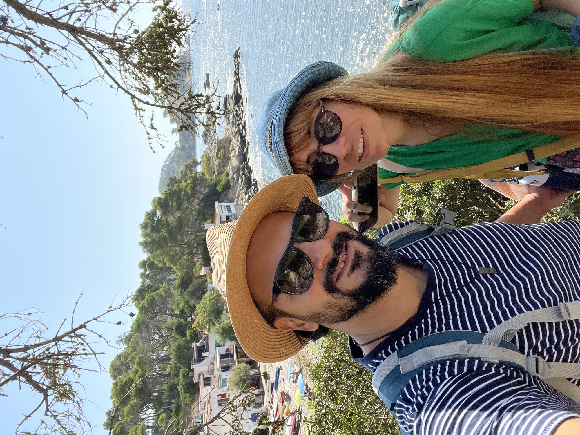 A man and a woman are taking a selfie outdoors with a body of water and a coastline in the background. The man is in the foreground on the left, wearing a striped t-shirt, a straw hat, and sunglasses. He has a beard and is smiling. He is carrying a backpack. The woman is on the right, wearing a green shirt and a blue bucket hat, and sunglasses. She is holding a phone, presumably to take the photo. Behind them, there are trees, white buildings with red roofs, a beach with colorful umbrellas and boats, and a rocky coastline leading to the sparkling blue sea under a clear sky. The scene appears to be a coastal area, possibly a small town or village, with lush vegetation and a clear, sunny day. The overall setting suggests a tourist destination, likely in the Mediterranean. The location is in the town of Palamós, Spain, which is known for its beautiful beaches and coastal scenery.