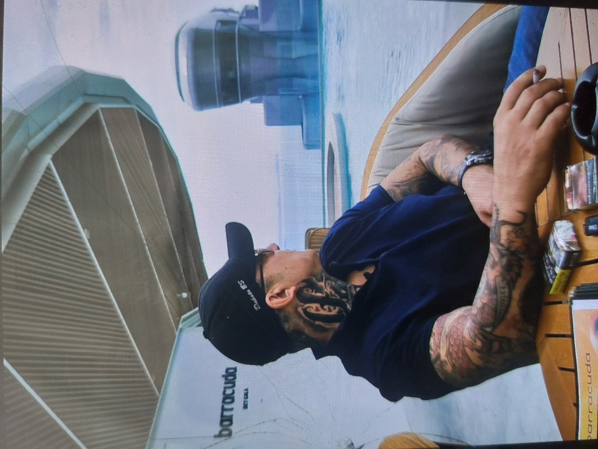A tattooed man wearing a black "Dublin 85" cap and dark blue shirt sits in a chair at what appears to be an outdoor cafe called "barracuda". He is holding a cigarette. The location is likely near water, given the background's reflective surface. The image was possibly taken in Tsarevo, Bulgaria, which has a "barracuda" restaurant.