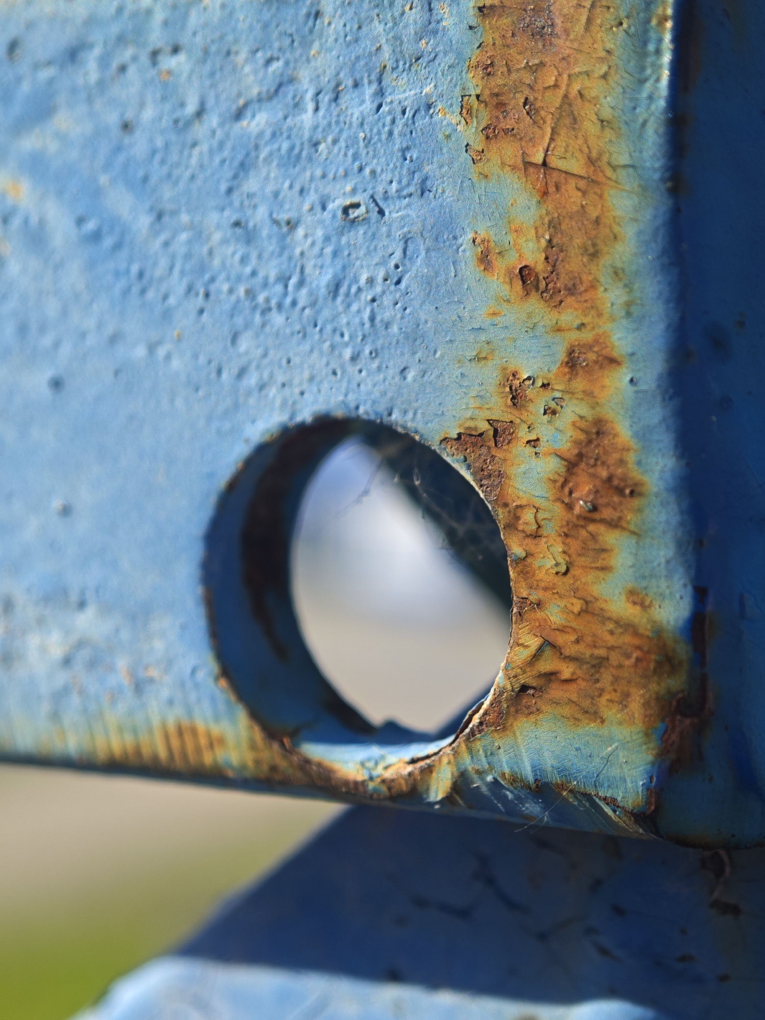 A close-up, macro shot captures a section of blue painted metal with significant rust. A circular hole is visible in the foreground, revealing a blurred background of light and a hint of green. The blue paint is textured with small indentations and shows signs of chipping. A large streak of orange and brown rust runs vertically along the right side of the frame, adhering to the metal's surface. The rust has a flaky, textured appearance, with some areas flaking away. The lighting suggests an outdoor environment, with bright light casting subtle shadows and highlighting the textures of the metal and rust. The overall impression is one of weathered material, with the blue paint struggling to protect the underlying metal from corrosion.