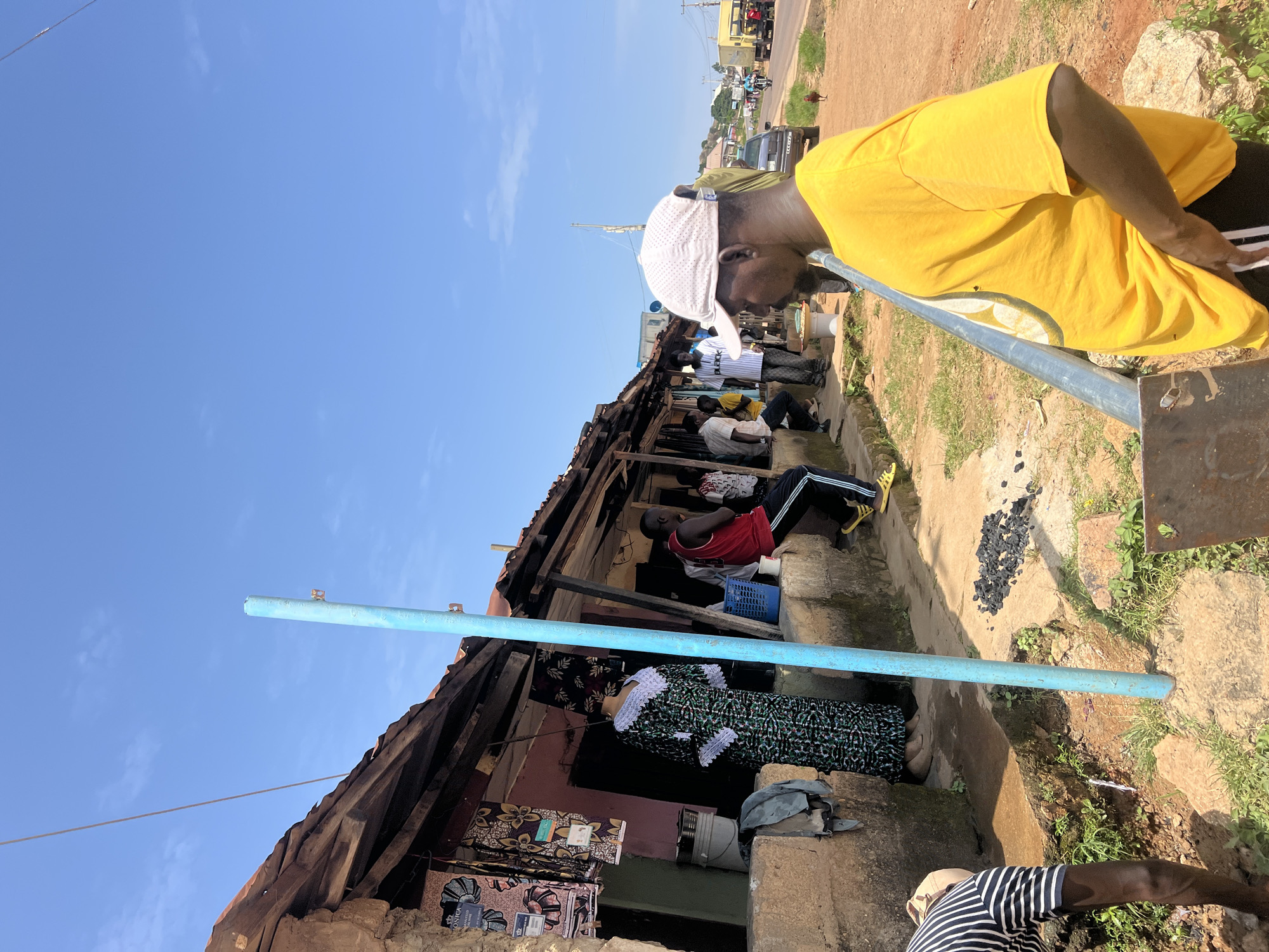 On a sunny day in Rukuba Road, Nigeria, a street scene unfolds with several people gathered near a row of buildings that appear to be shops or stalls. In the foreground, a person wearing a yellow t-shirt and a light-colored baseball cap faces away from the camera, with their right hand in their pocket. To the left, another individual wearing a striped t-shirt and dark pants is partially visible, appearing to be engaged in some activity.
Behind them, a woman in a patterned dress stands near a display of colorful fabrics hanging outside one of the establishments. Several men are seated or standing along the front of the buildings. One man in a red tank top and black pants with white stripes sits on a ledge. Another man in a white shirt and dark pants stands further down the row.
The buildings have open fronts and covered porches, with corrugated metal roofs. A blue pole stands prominently in front of the shops. In the background, a road is visible with vehicles, including a yellow truck and a car, suggesting a commercial or transit area. The ground is a mix of dirt and scattered charcoal. The sky is clear and blue.