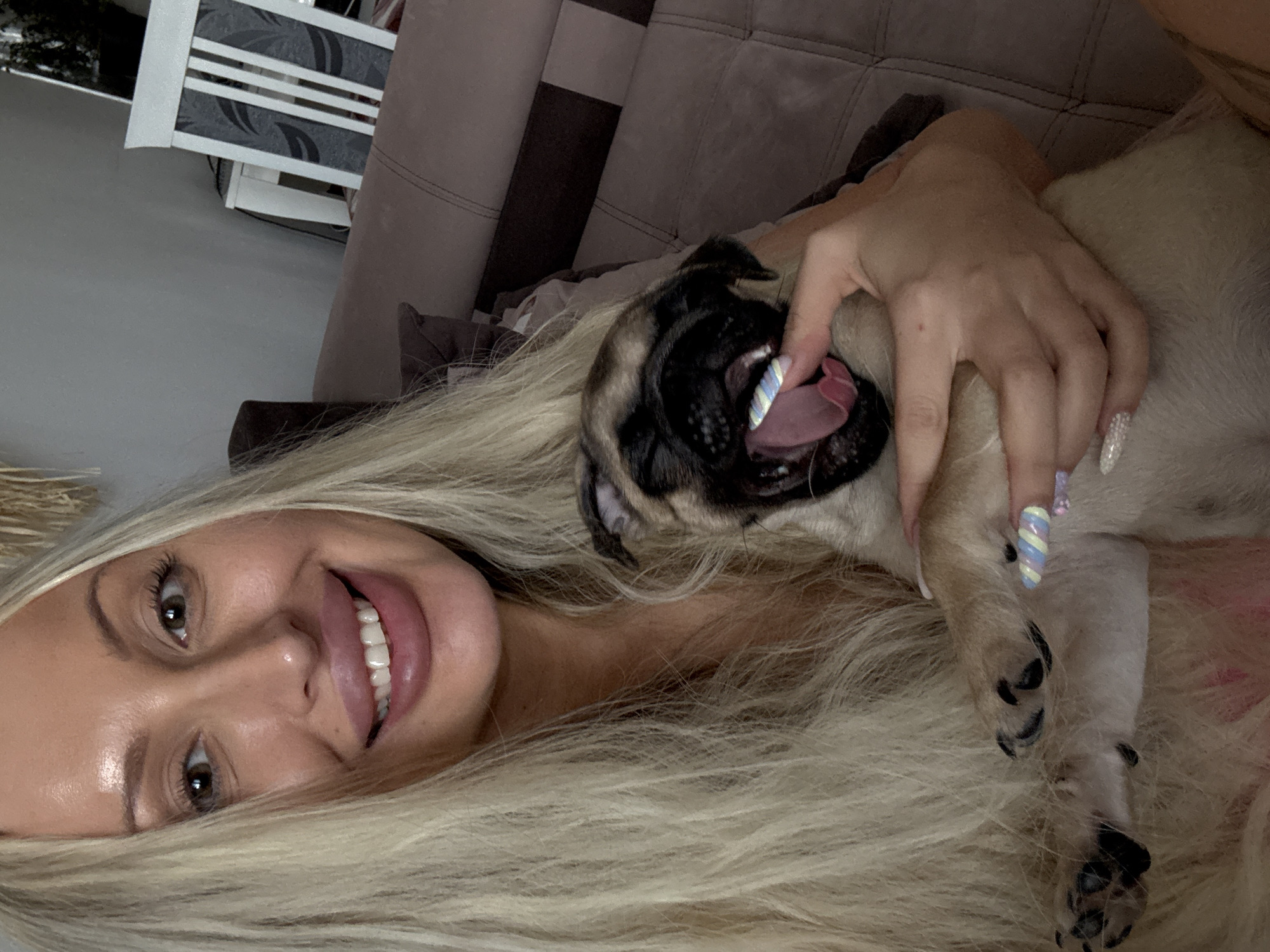 A blonde woman with full lips and long blonde hair is smiling at the camera while holding a pug. The pug is yawning with its tongue out. The woman's nails are painted with pastel colors and glitter. They are sitting on a brown sofa in an indoor setting. A white chair with a dark pattern is visible in the background.