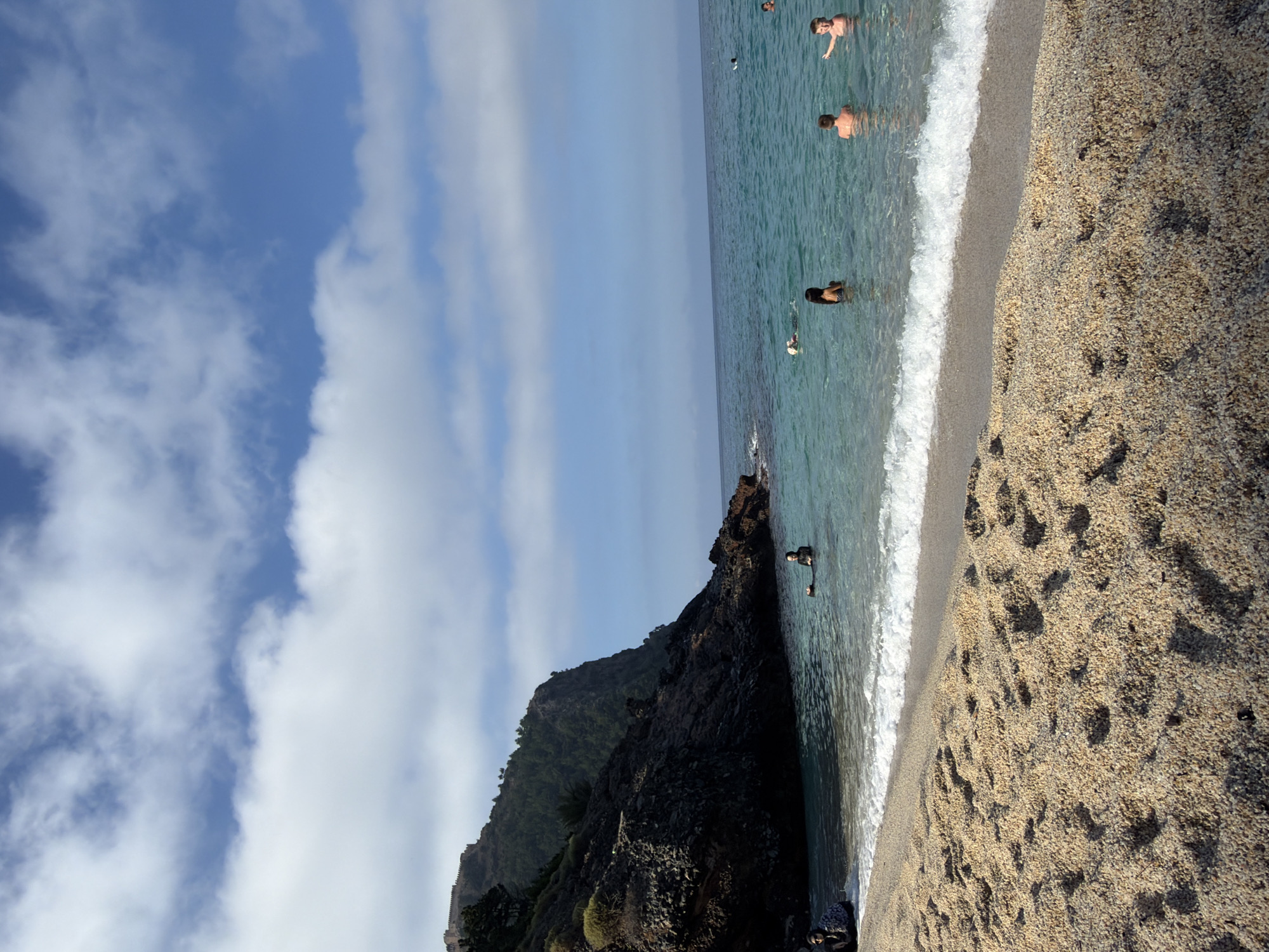 The image depicts a sunny day at a beach with a rocky cliff face on the left. The water is clear and turquoise, with several people swimming and wading. Some individuals are visible in the shallow water near the shore, while others are further out. The beach itself is sandy with some pebbles. Above the cliff, a structure resembling a castle or ancient fortification can be seen. The sky is blue with scattered white clouds.