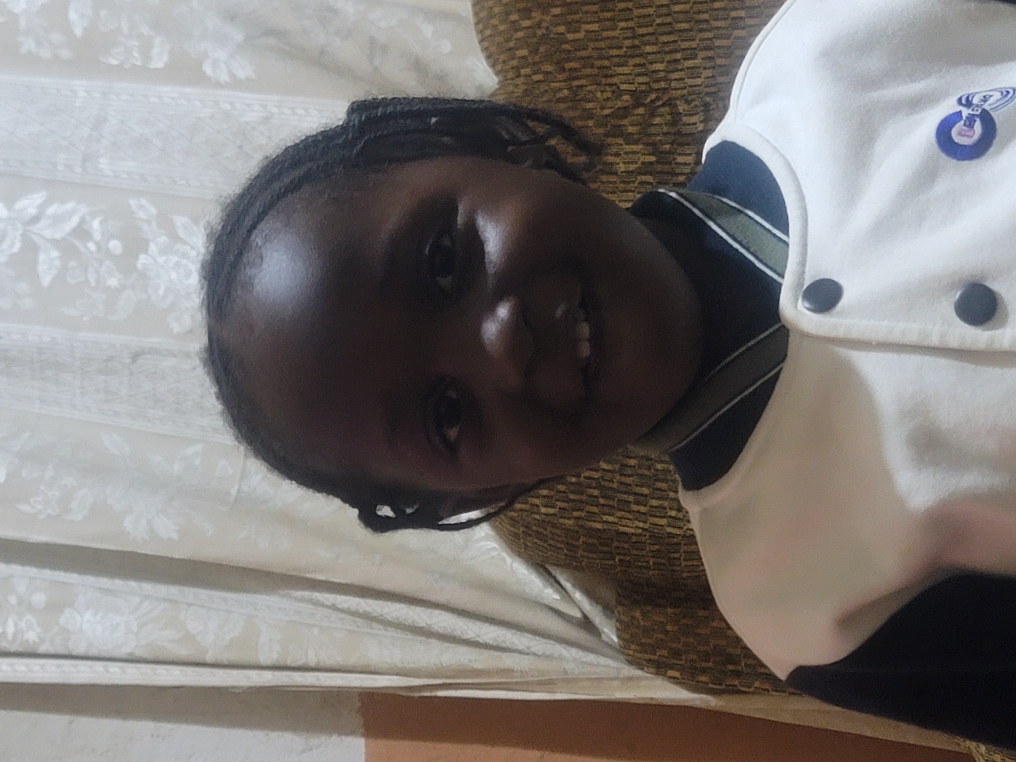 A frontal portrait captures a young child with dark skin tone, seated indoors in the city of Unknown, Unknown. The child faces directly forward, looking at the camera with wide eyes and a faint smile, revealing some upper teeth. Their dark hair is styled in multiple braids pulled away from the face. They are wearing a white, long-sleeved jacket with dark (likely navy blue) sleeves. The jacket features a ribbed collar in a similar dark color, accented with two light-colored stripes. Two dark circular buttons are visible down the front placket. A blue circular logo with white abstract elements is present on the right chest area of the jacket. The background consists of light-colored (cream/off-white) curtains with a subtle floral or leaf pattern, displaying vertical folds. Behind the child's shoulders, a section of upholstered furniture with a textured, woven pattern in shades of brown and possibly gold is visible. A sliver of a light brown/orange wall is present on the far left.
