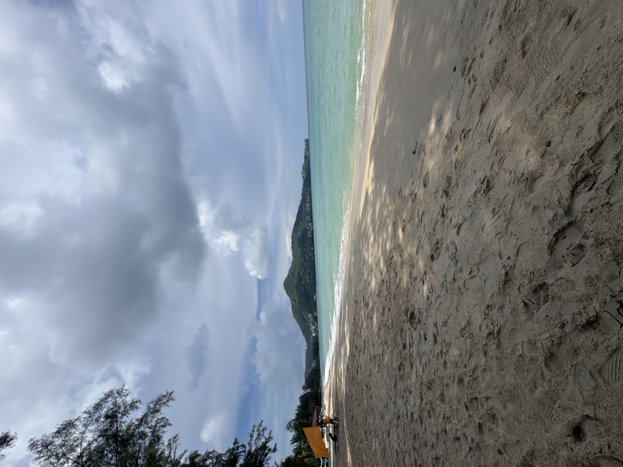 The image shows a wide, sandy beach with gentle waves lapping the shore under a cloudy sky. In the foreground, the sand is textured with footprints and debris, cast in dappled sunlight. To the left, a bright orange umbrella provides shade for two individuals, one sitting and one standing. Behind them, trees line the edge of the beach, and further in the distance, a lush green hillside dotted with buildings slopes down to the turquoise ocean. The scene suggests a serene, tropical atmosphere in Ban Bang Thao, Thailand.