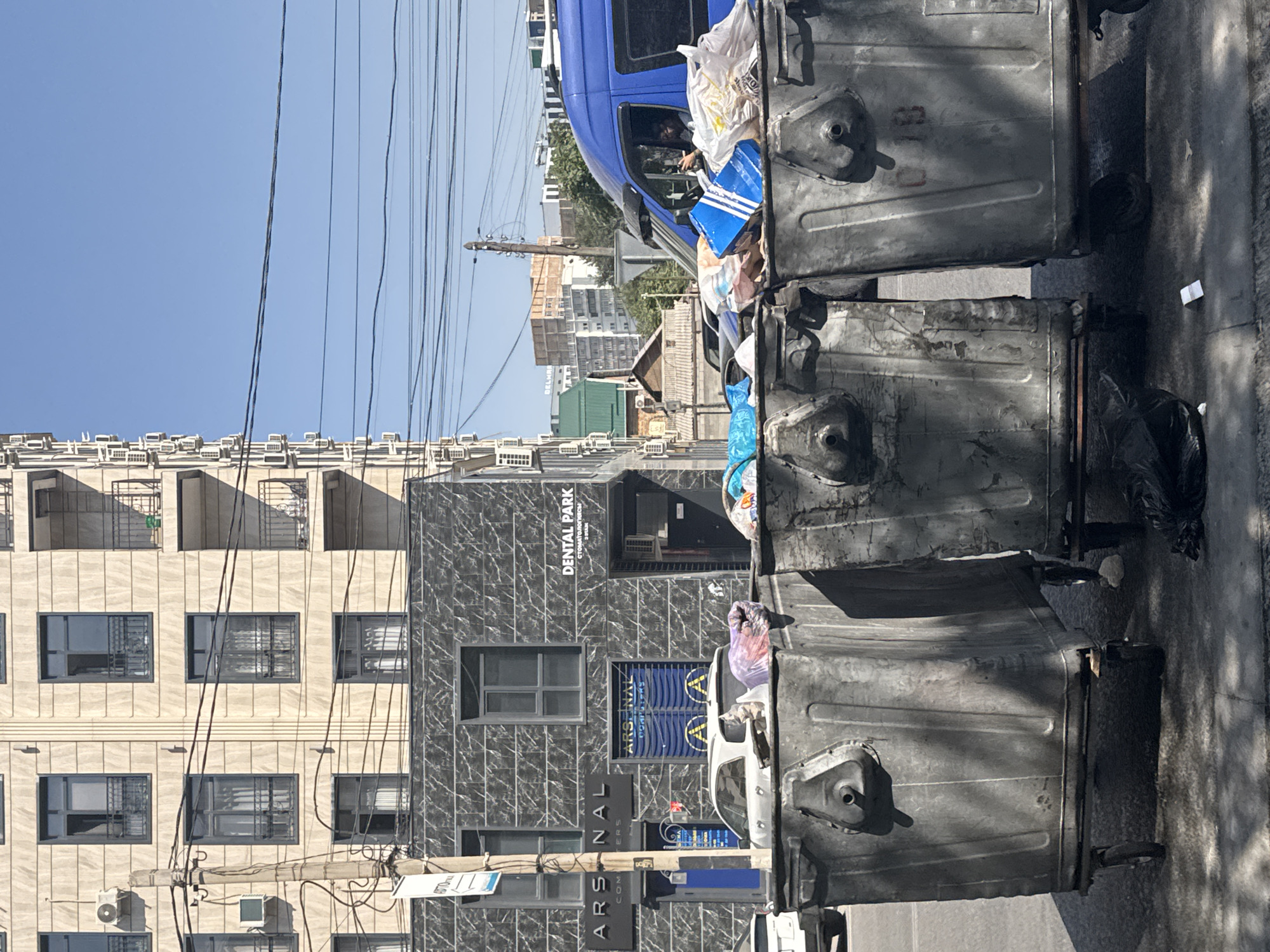 The image shows a street scene in Bishkek, Kyrgyzstan, featuring a row of overflowing trash bins in the foreground. Behind the bins, a modern building with a dark, patterned facade stands. Signs on this building indicate "ARSENAL COMPUTERS" and "DENTAL PARK". Several cars are visible parked in front of the building, and a blue van is parked on the right side of the image, with a person visible inside, using a mobile phone. The background shows a blue sky with power lines stretching across it, and other urban buildings. The trash bins themselves are large, metal containers, some with lids ajar, revealing the contents within, which include plastic bags and general refuse. The overall impression is one of urban life with a focus on public waste management.
