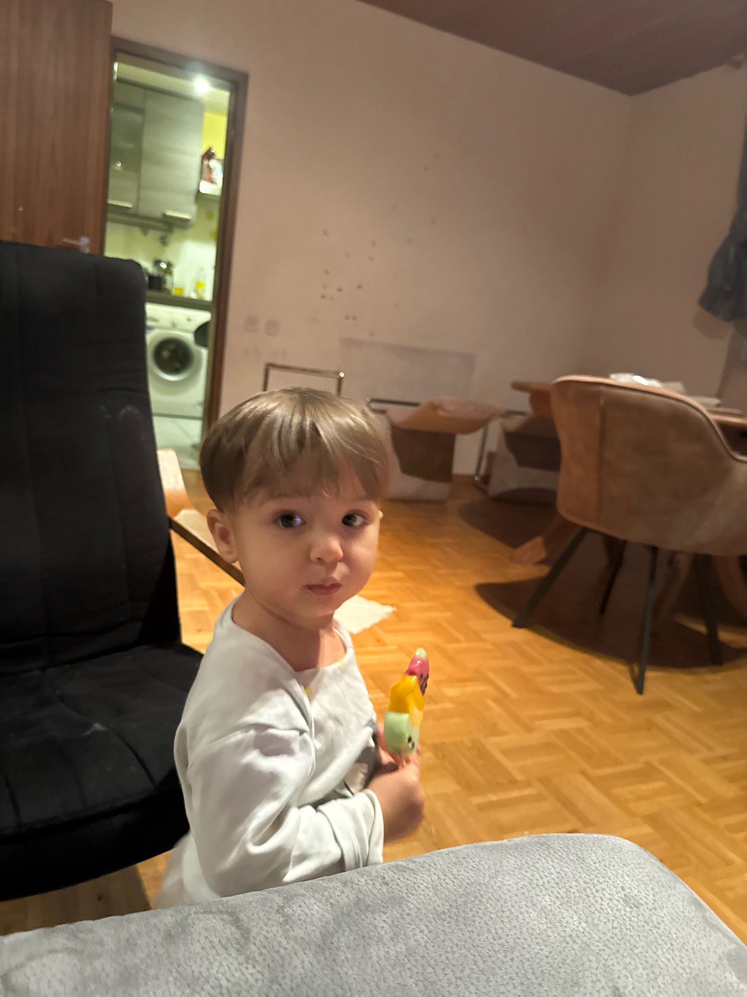 In Überherrn, Germany, an indoor scene is depicted. A young child with light brown hair is positioned centrally, holding a multi-colored popsicle. The child wears a long-sleeved white shirt. To the left of the child is a large black chair. In the background, through an open doorway, a kitchen area is visible, featuring a washing machine and cabinetry. The floor is made of polished wood. Several dining chairs and tables are present in the middle ground and background, suggesting a dining or living area.