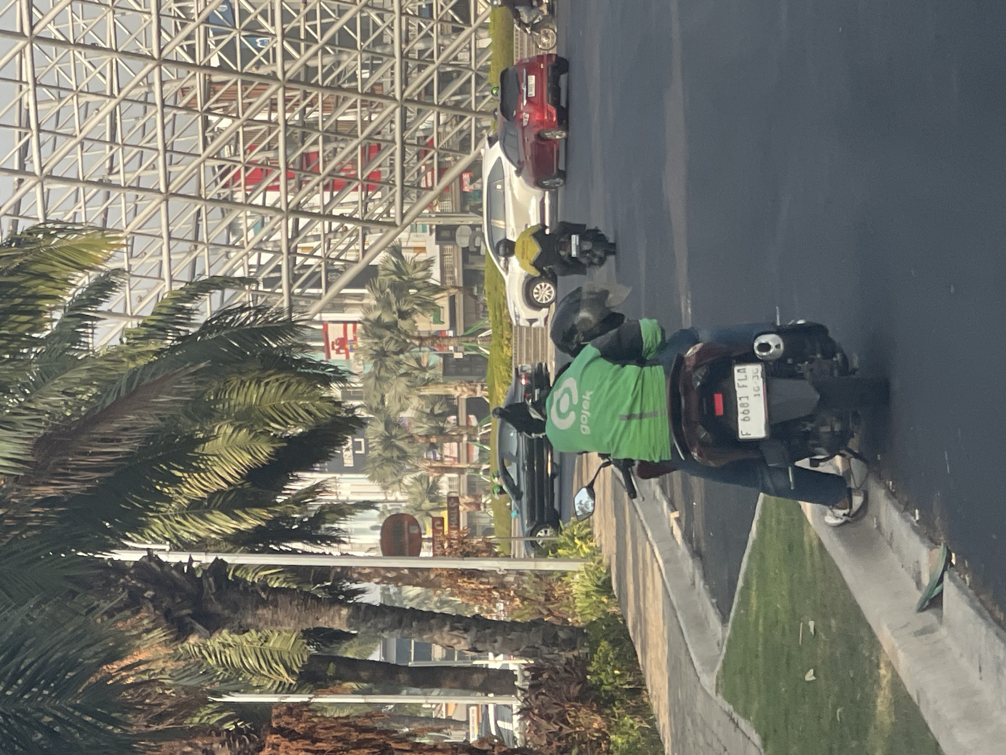The image shows a street scene in Bekasi, Indonesia. In the foreground, a person on a motorcycle wears a bright green jacket with the Gojek logo. The motorcycle has a license plate that reads "F 6681 FLA 10-26". The person is wearing blue jeans and white shoes. In the background, several cars are visible, including a black sedan, a white SUV, and a red hatchback. Palm trees line the street, and a modern building with a steel lattice structure is prominent in the distance. The environment appears to be urban, with a clear sky and daylight.
