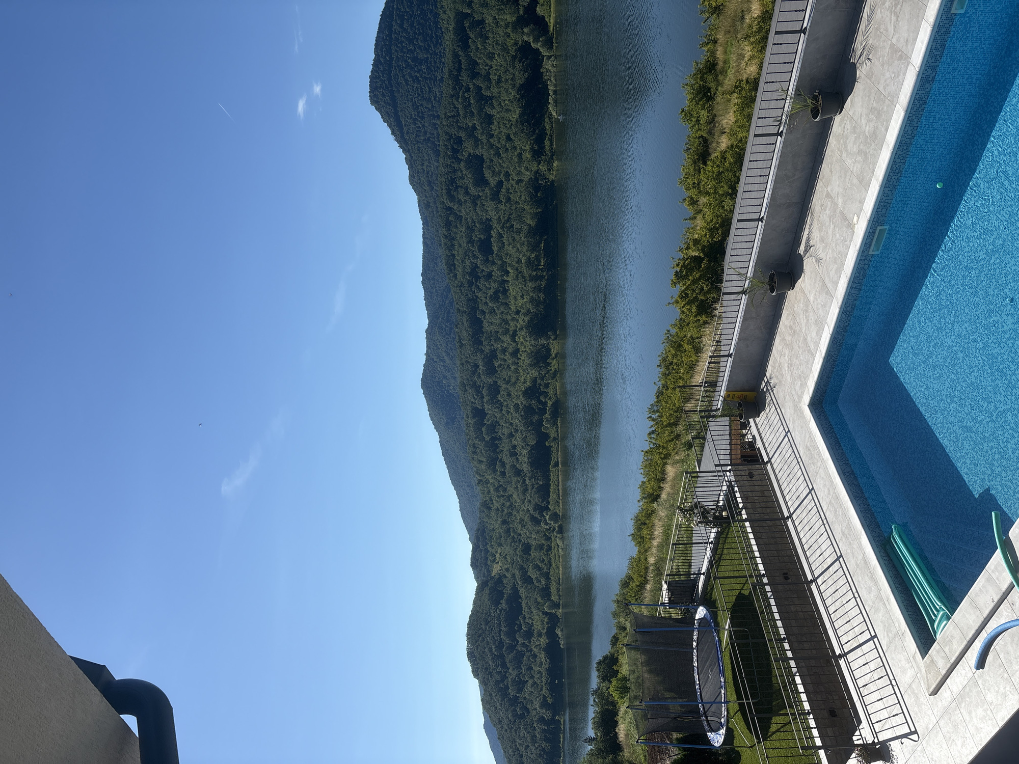 This is a high-angle, full shot image of a residential property with a swimming pool overlooking a body of water and forested hills. The foreground features a bright blue swimming pool with a green pool float. The pool deck is paved with light-colored tiles and lined with a dark metal fence. To the left of the pool, a trampoline with a black net is visible on a grassy area. A dark metal fence encloses the trampoline area. Beyond the fence, a vast body of dark blue water stretches into the distance, bordered by lush green trees that cover rolling hills. The sky is clear and bright blue. A gutter is visible in the upper left corner of the image, suggesting the viewpoint is from a balcony or roof. The scene appears to be a serene, outdoor recreational area in a natural setting.