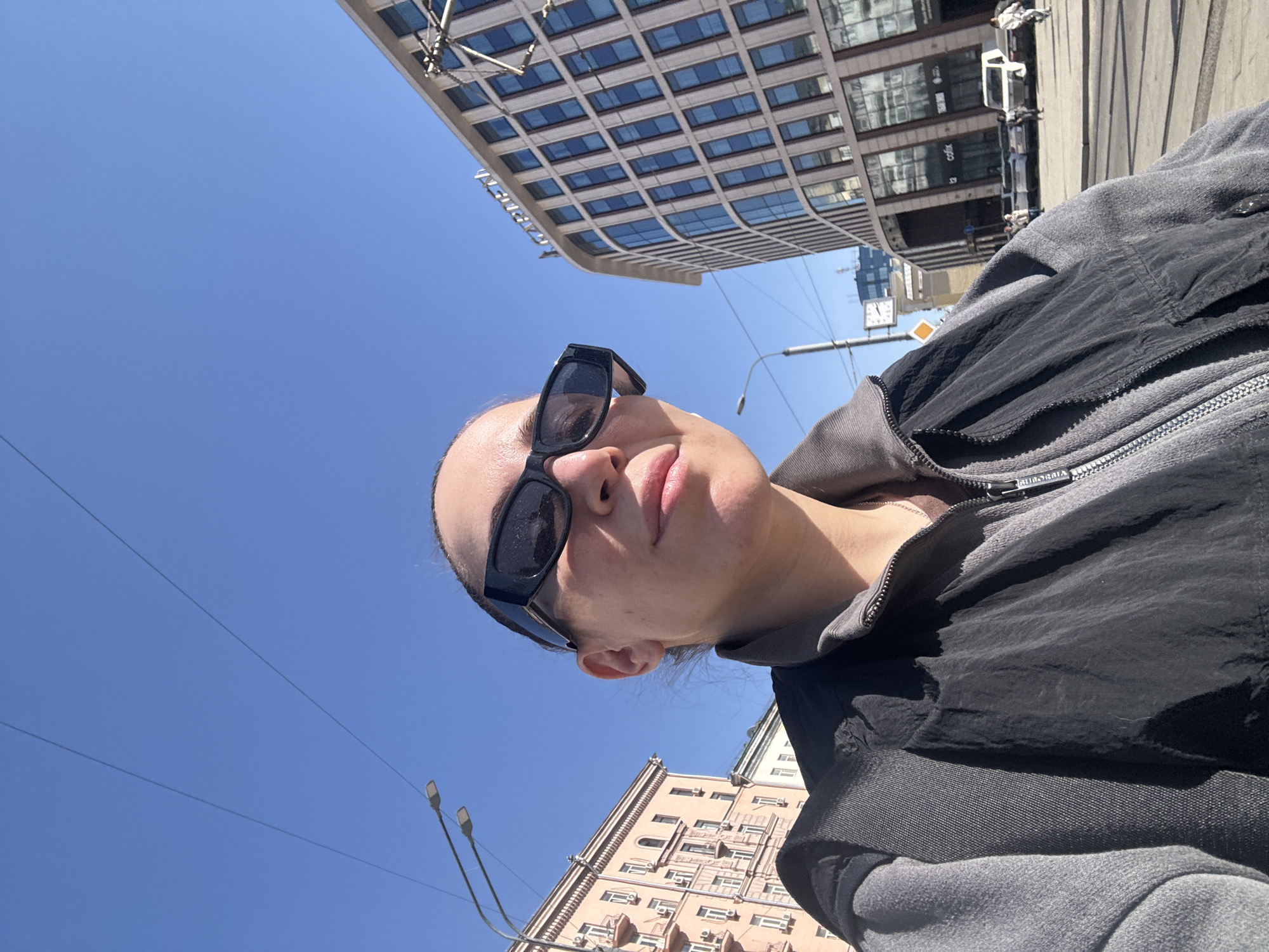 A low-angle, outdoor photograph shows a woman wearing sunglasses and a gray and black jacket, standing on a street in Moscow, Russia. The woman's head is tilted back, and she is looking slightly to the left. The sky is blue and clear. In the background, there are tall buildings, including one with a distinctive curved facade and numerous windows, and another that is pink. Streetlights and power lines are visible against the blue sky. A clock is visible on one of the buildings.