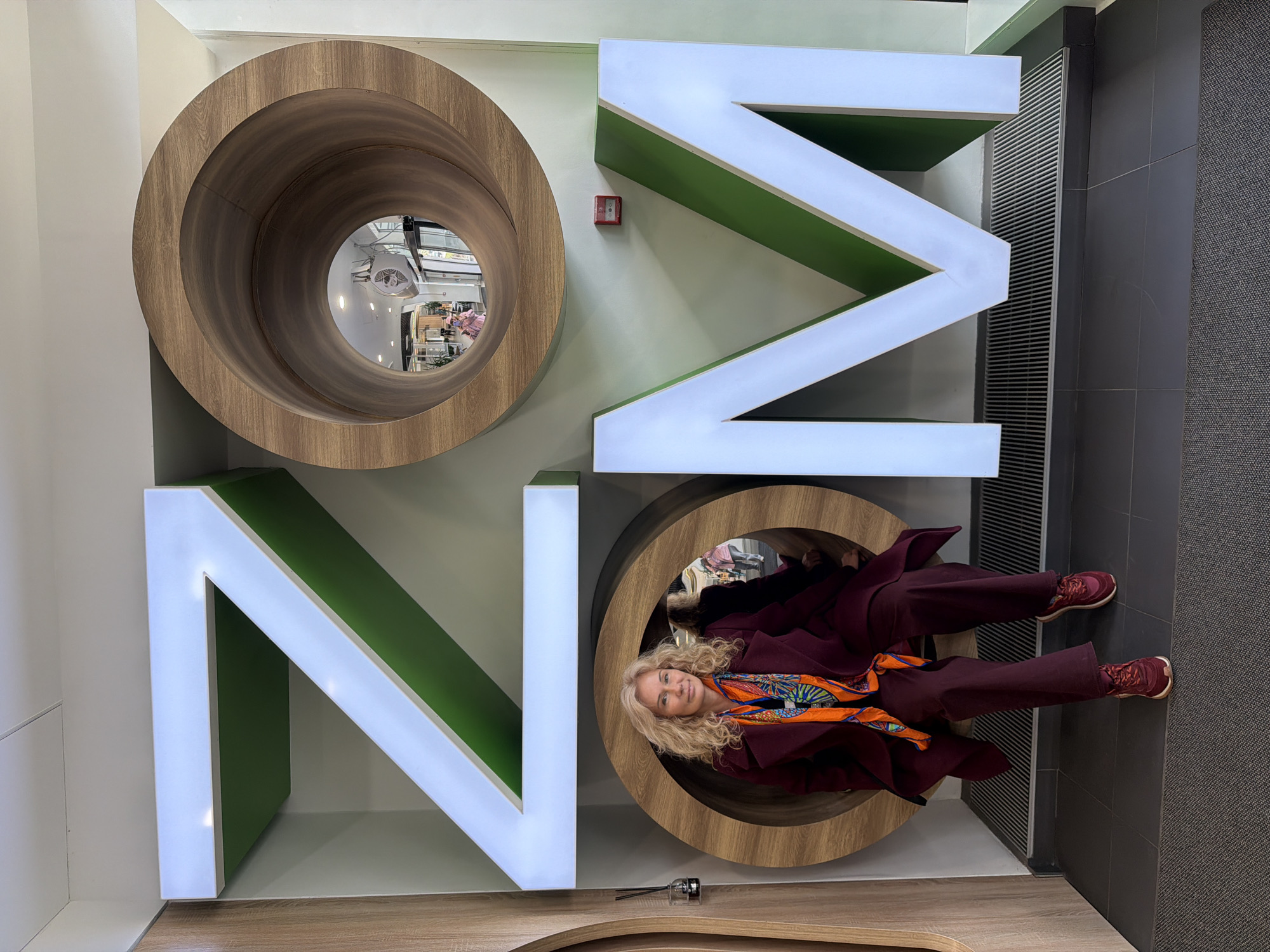 The image features a woman sitting inside the letter "O" of the word "ON", which is constructed from large wooden and white letterforms. The letters are mounted on a wall with the letters oriented to spell "ON" when viewed from the side. The "O" is wooden and cylindrical, and the "N" is white with a green interior.
The woman has blonde curly hair and is wearing a maroon coat, matching maroon pants, an orange patterned scarf, and maroon sneakers. She is positioned inside the bottom "O". The top "O" is a cylinder with a reflective interior, suggesting a mirror or a glossy surface.
The background includes parts of the wall and ceiling, which are light green, and a section of the floor, which has a wooden finish. A small red alarm box is visible on the wall between the letters. The composition and orientation of the image suggest a playful or artistic installation.